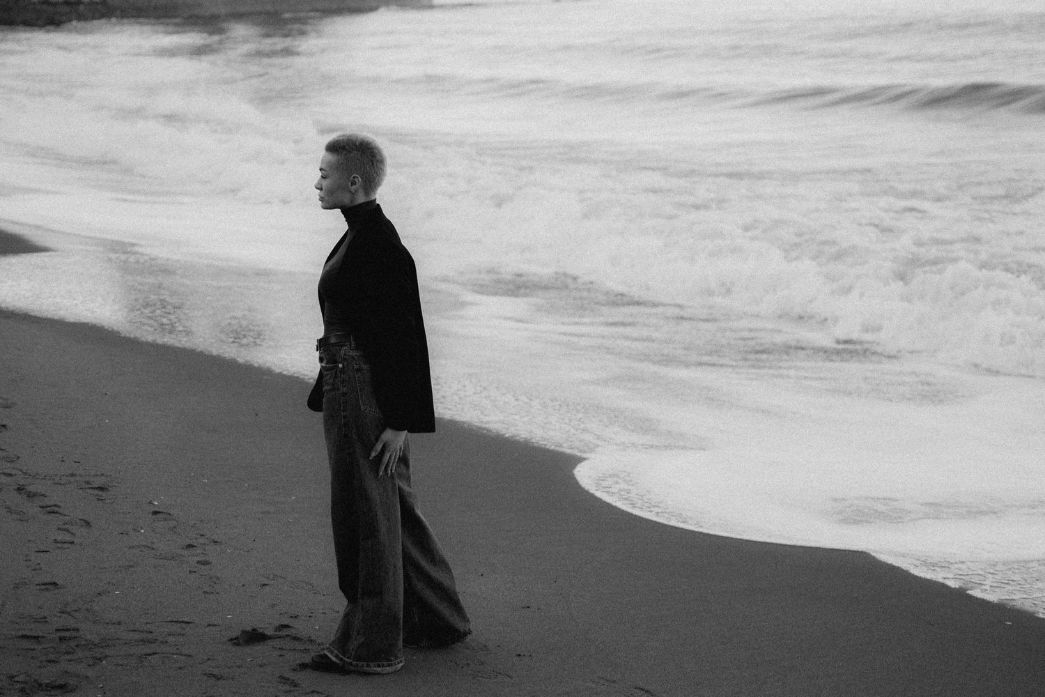 A silhouetted figure walks along a misty beach shoreline at dusk as waves roll onto the sand.