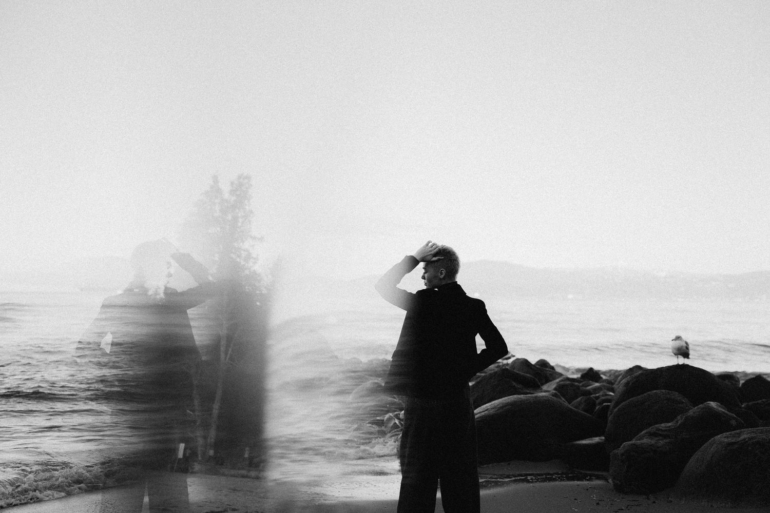 Silhouetted figure stands on rocky coast as waves crash and spray against dark shoreline at sunset.