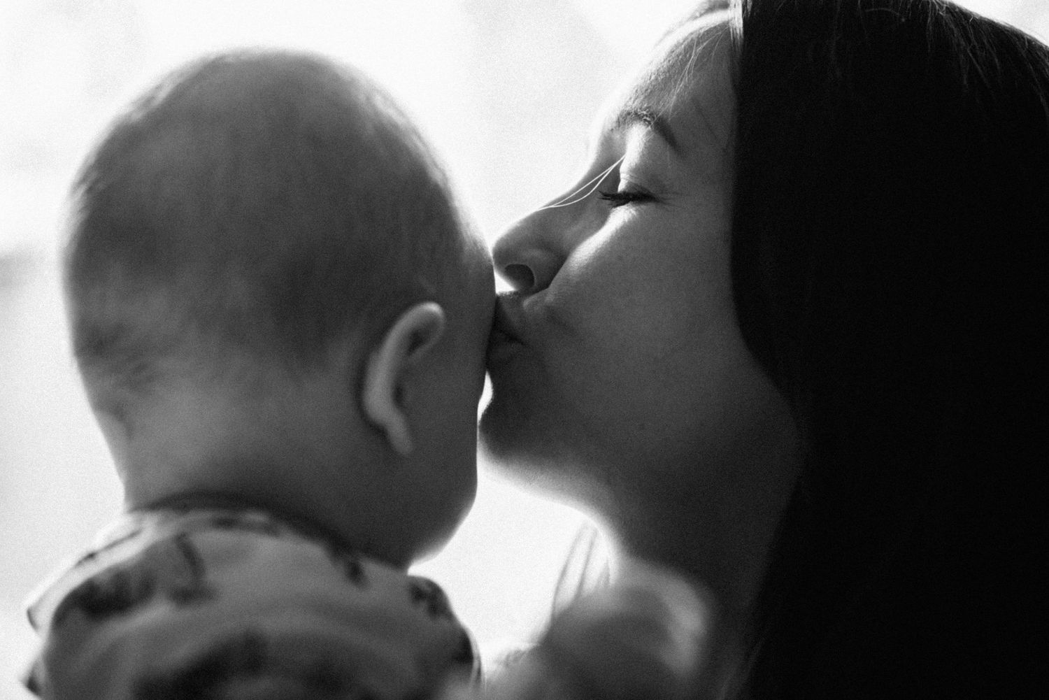 A tender black and white portrait of a mother kissing her baby's head in soft focus lighting.