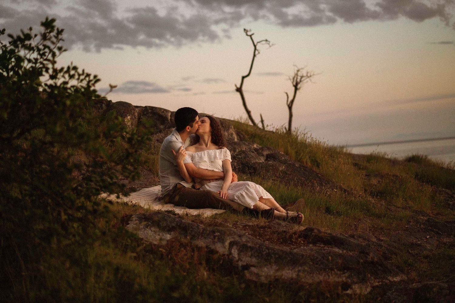 A romantic sunset portrait of two people sitting on a hillside overlooking the water.