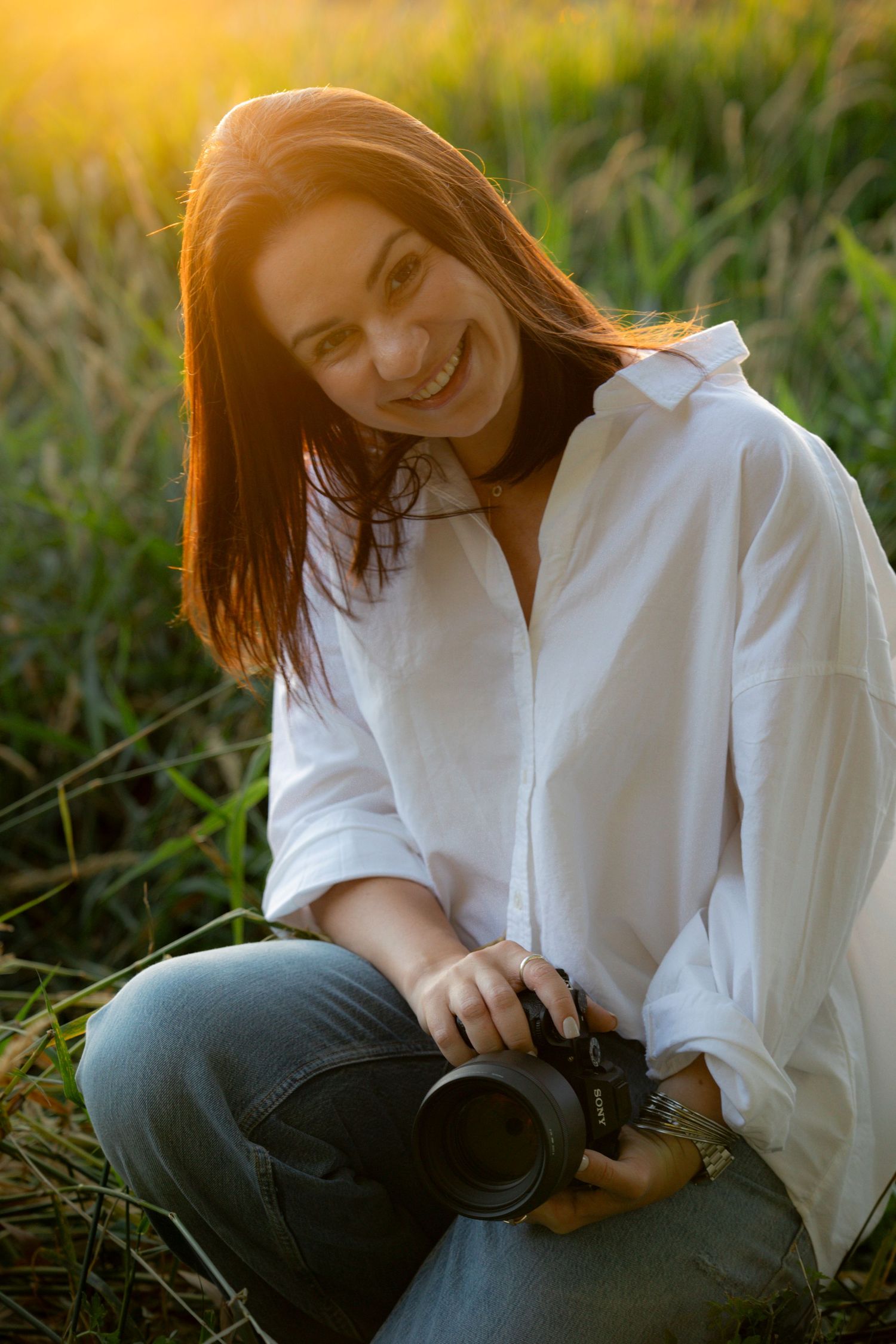 A photographer in a white shirt smiles while holding camera equipment in golden grass.
