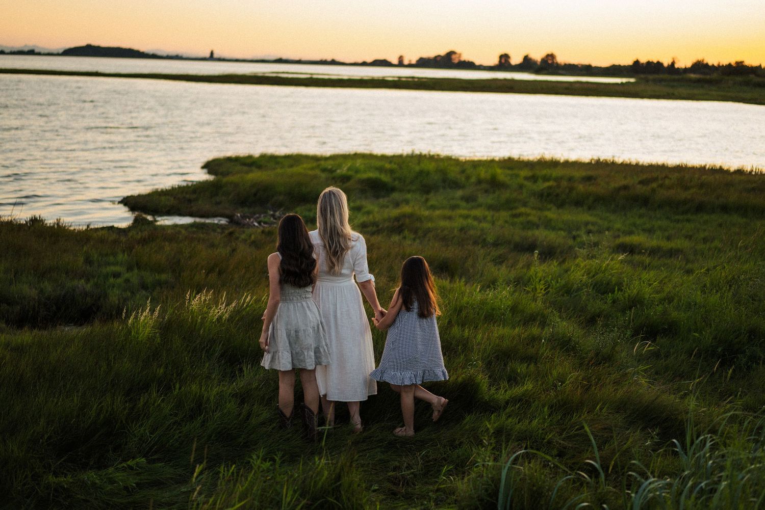 Three figures in white dresses stand together watching the sunset over a serene waterfront landscape.