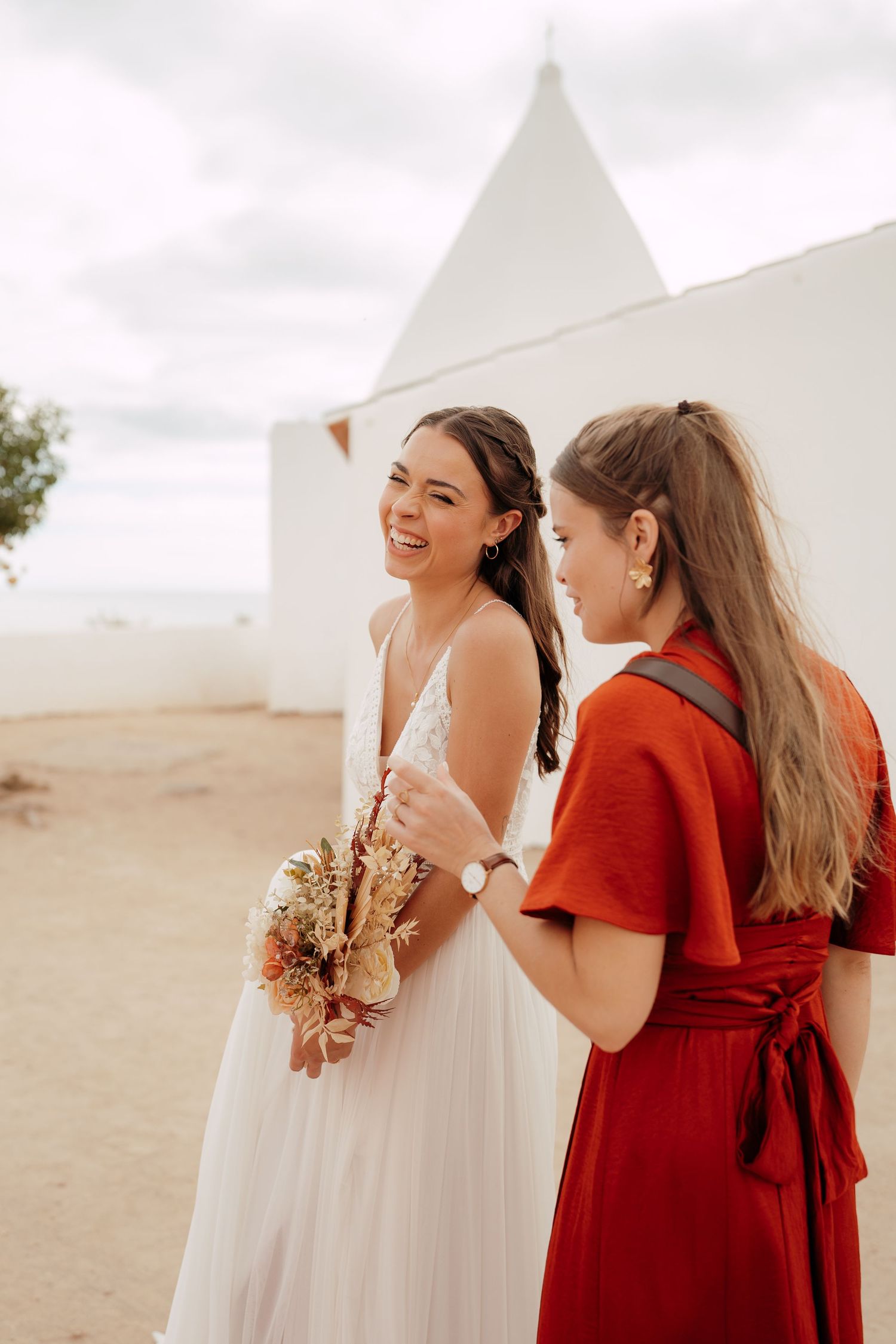 Smiling bride in white dress holding bouquet stands with bridesmaid in red dress near white chapel.
