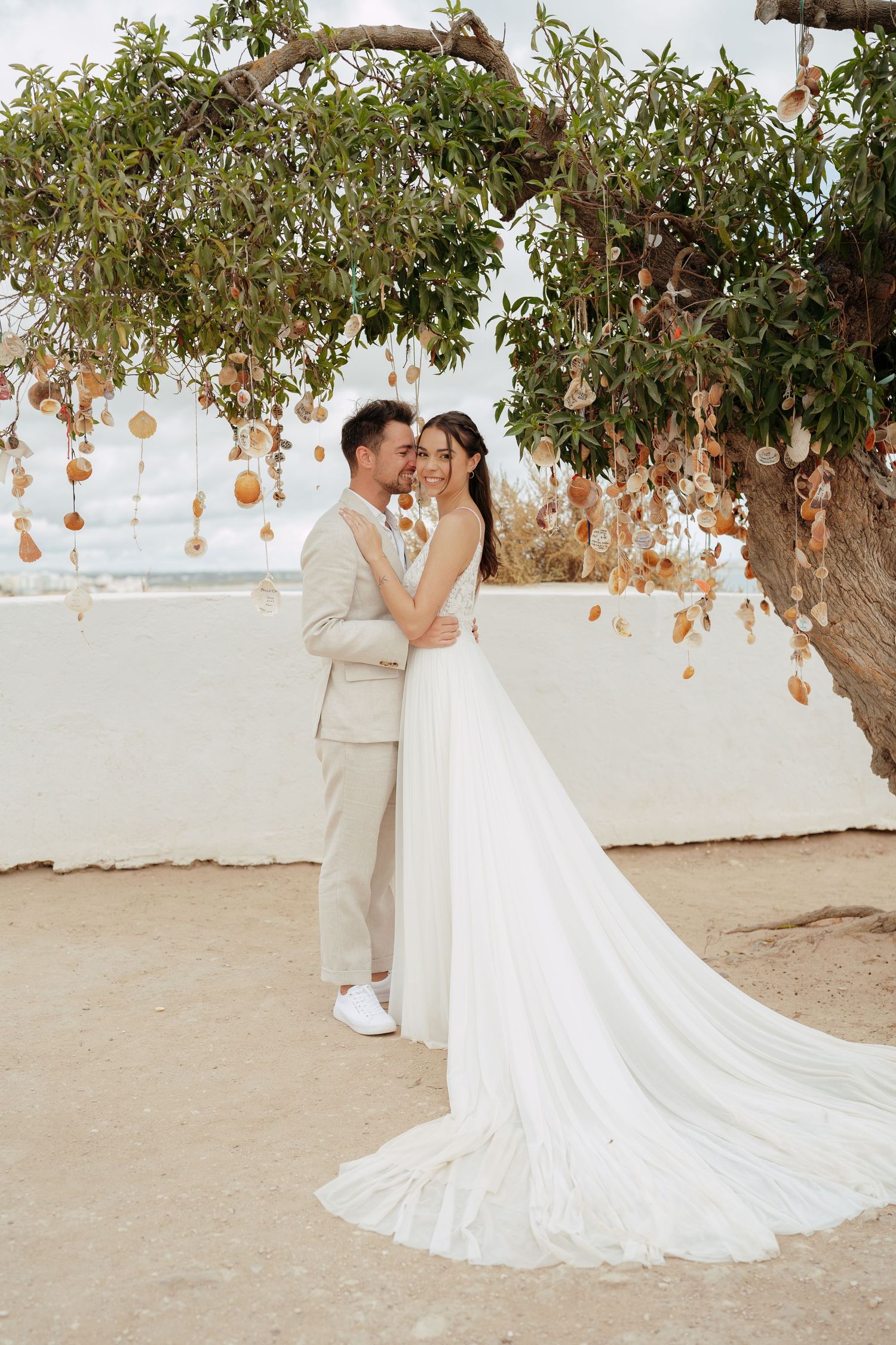 Bride and groom embrace under a decorated tree with hanging ornaments at an outdoor beach wedding ceremony.