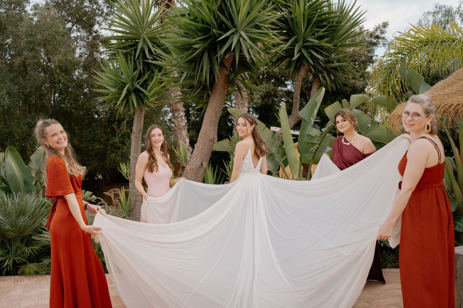 Bride and bridesmaids in red and pink dresses holding flowing white bridal gown in lush tropical garden.