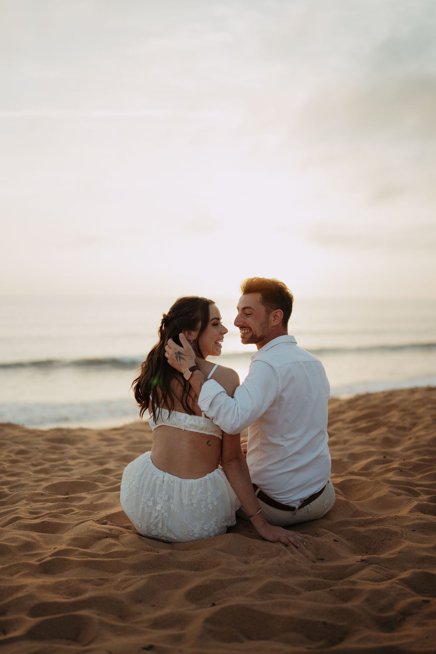A couple sits romantically on sandy beach at sunset, the man in white shirt embracing the woman in a white outfit.