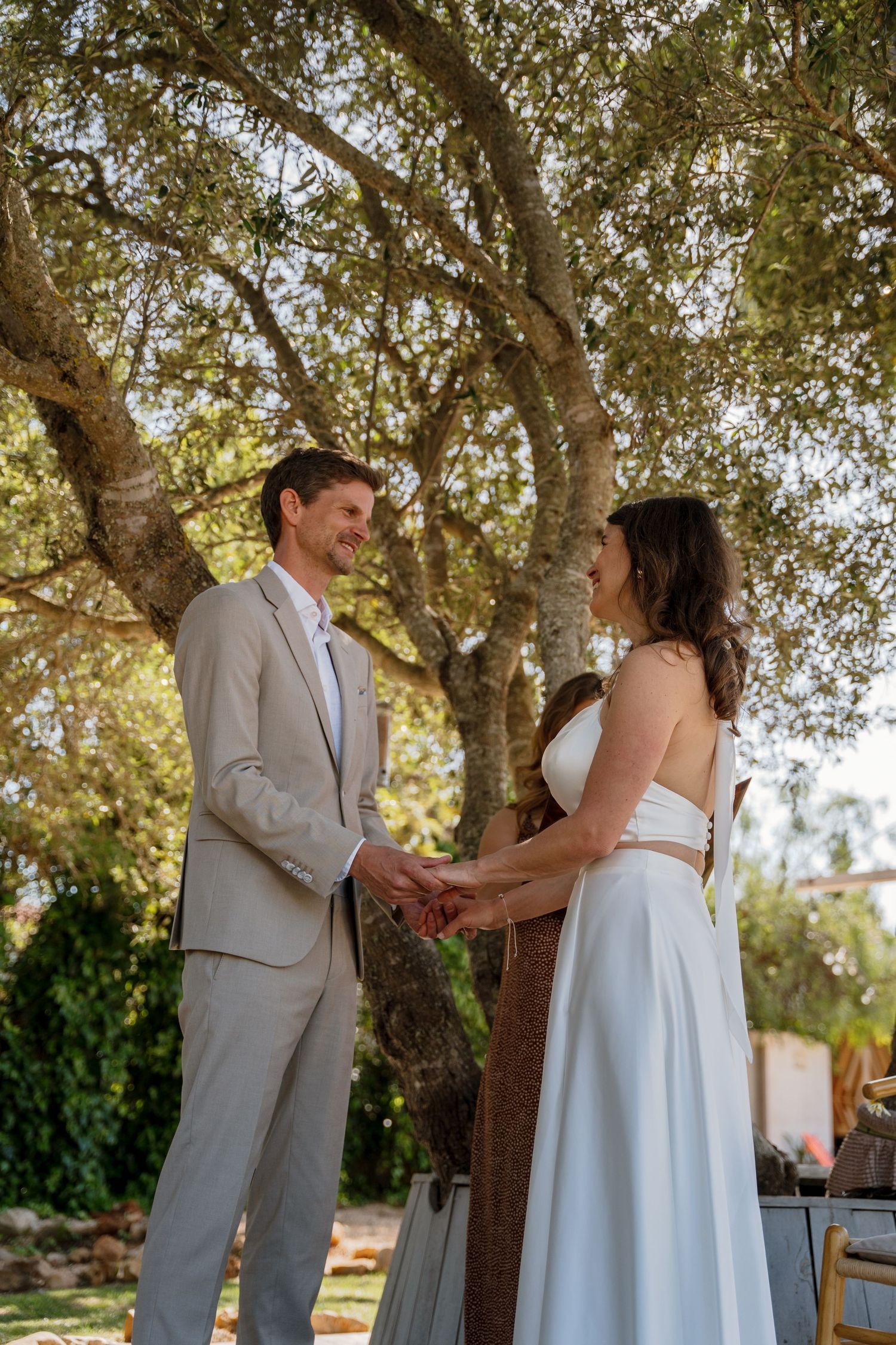 Smiling couple holds hands while exchanging wedding vows beneath a large olive tree outdoors.
