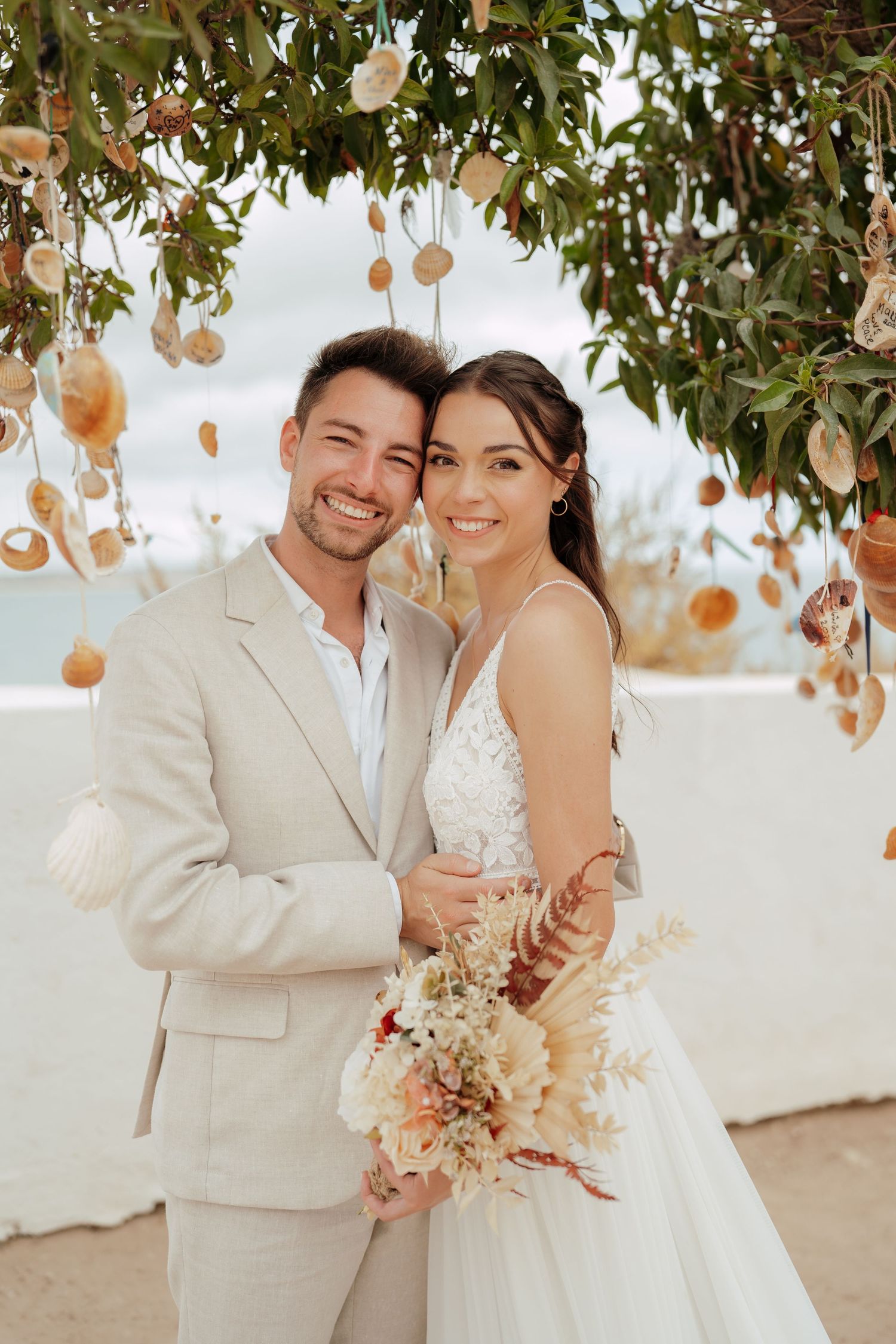 Smiling bride and groom pose under seashell-adorned tree arch at beach wedding, bride holding boho floral bouquet.