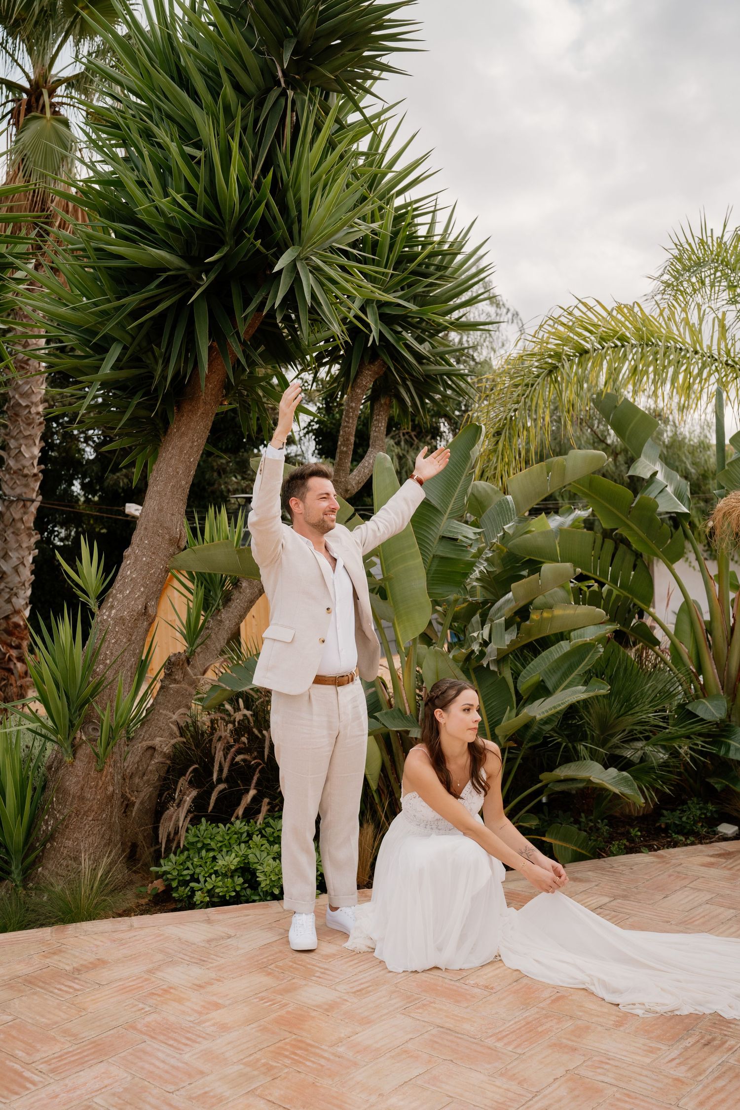 Bride in white gown sits on terrace while groom in beige suit raises arms among tropical palms.