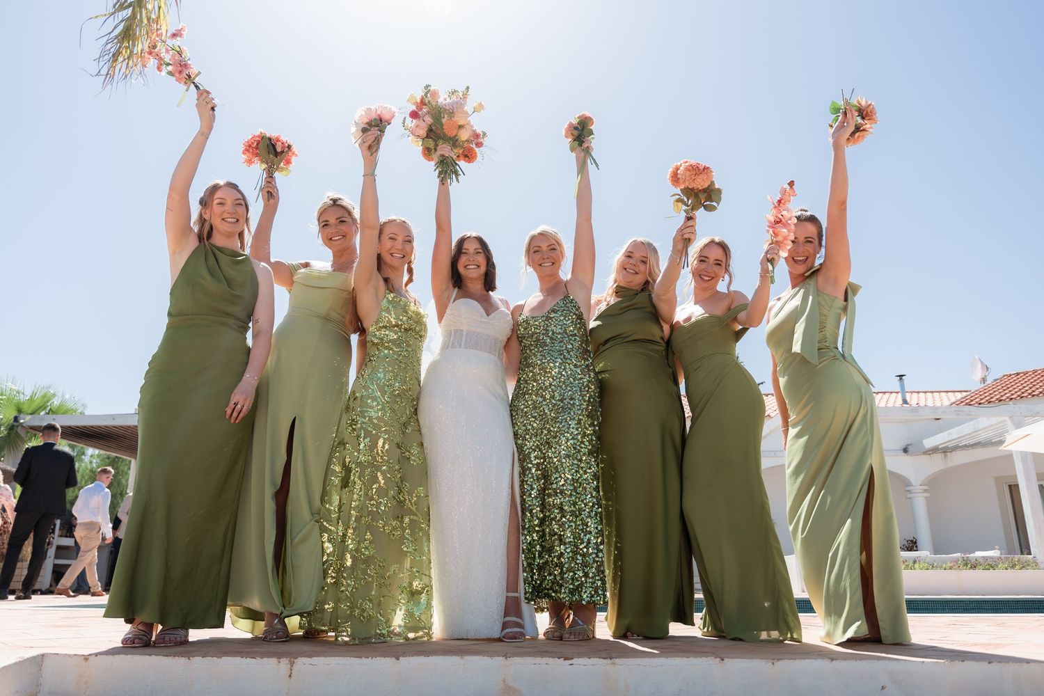 Bride in white gown with bridesmaids in olive green dresses raising bouquets outdoors on sunny day.