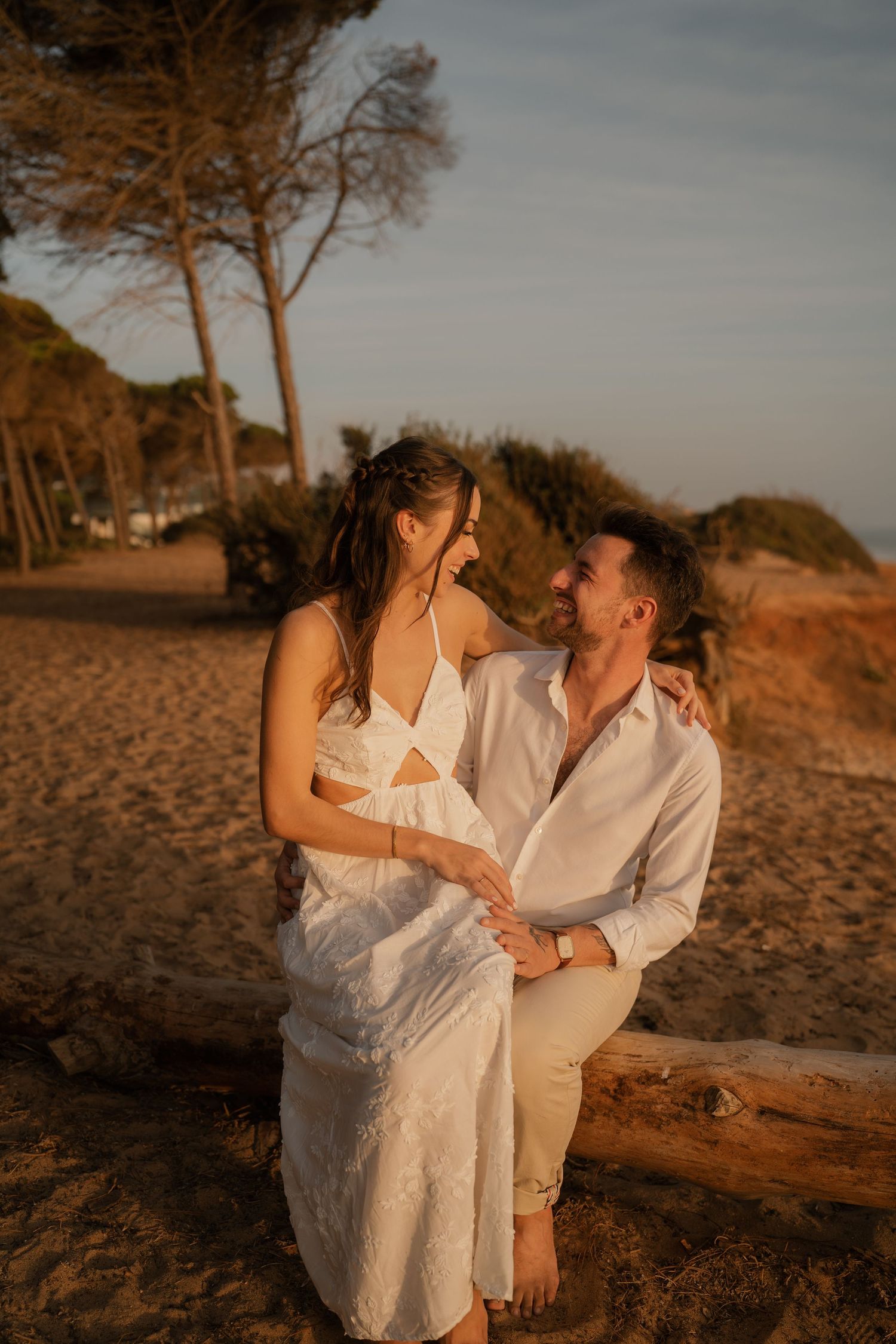 Couple in white outfits sharing a romantic moment on a sandy beach at golden hour, seated on driftwood.