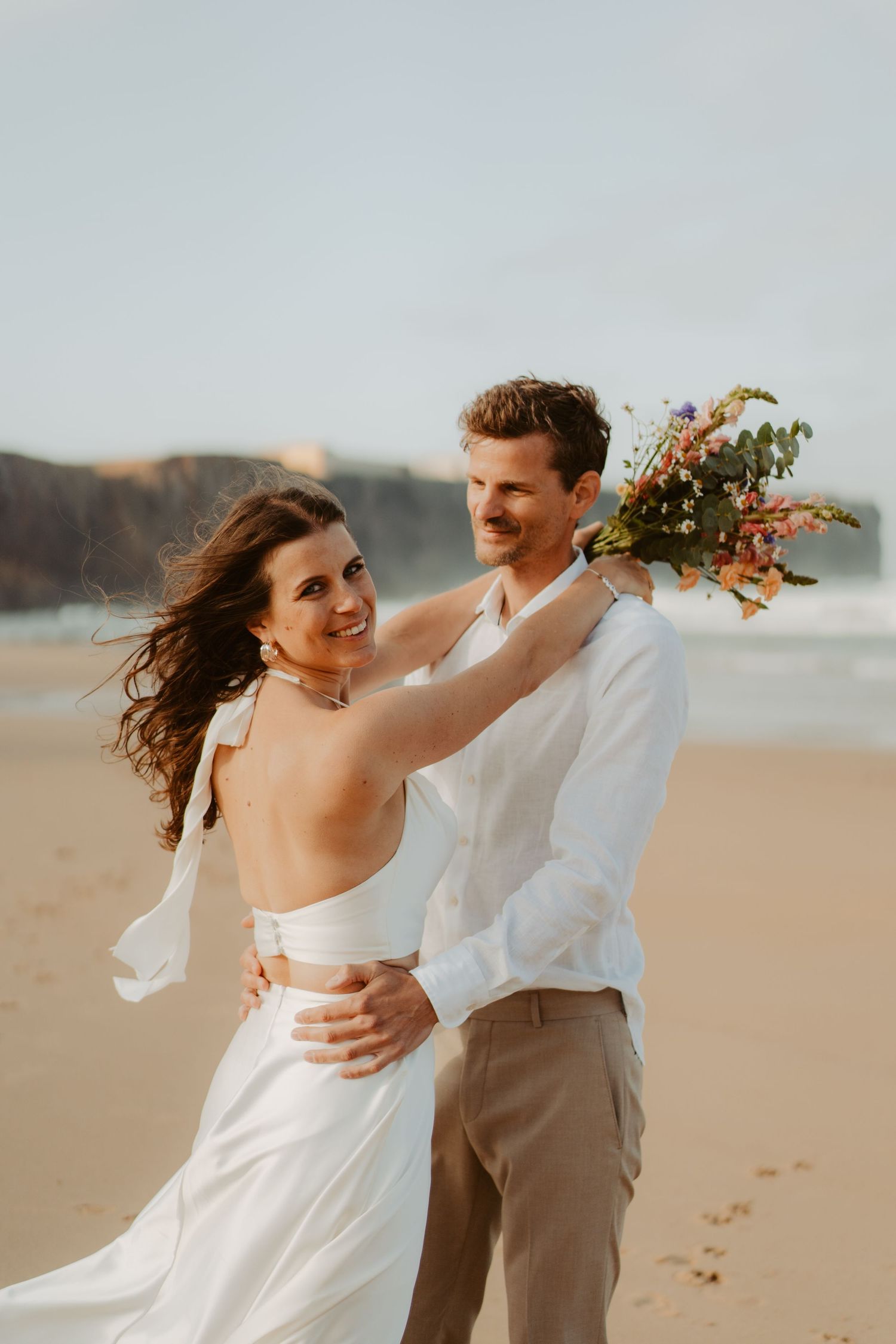 Bride rests arm on grooms shoulder holding bouquet as they smile together on a sunny beach.