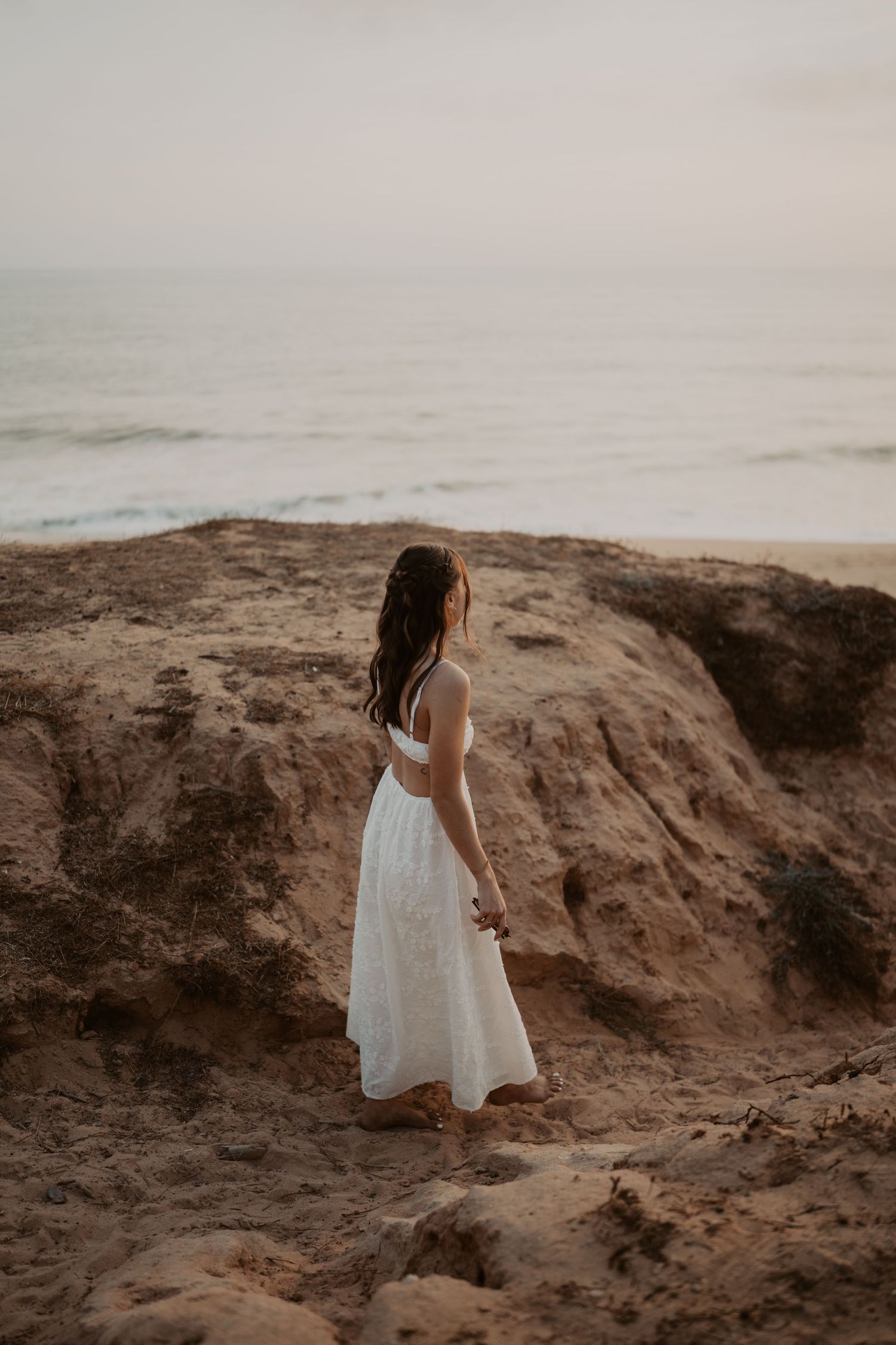 Woman in white dress standing on sandy cliffs overlooking the ocean at dusk.