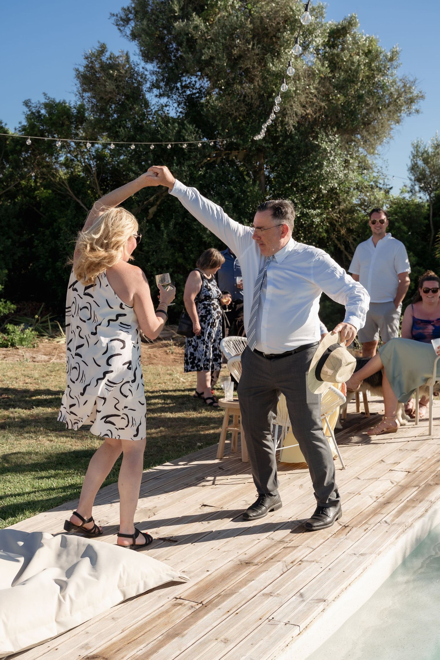 Couple dancing outdoors on wooden deck at garden party near pool, guests watching in background on sunny day.