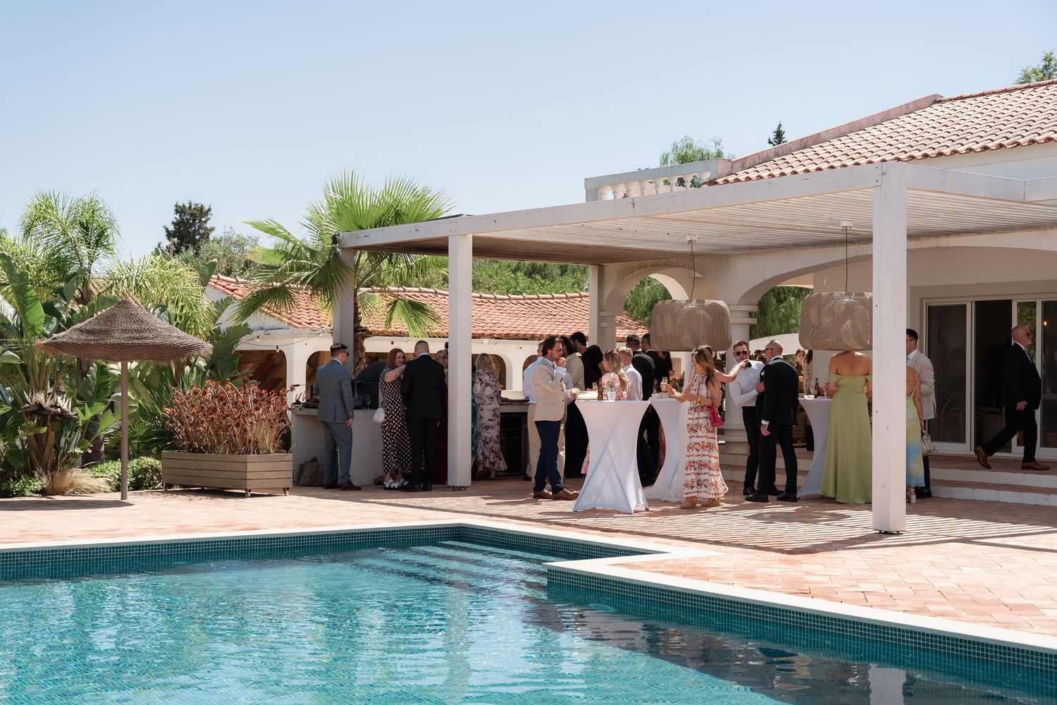 Elegant outdoor poolside reception with guests mingling under a white pergola at a luxury villa.