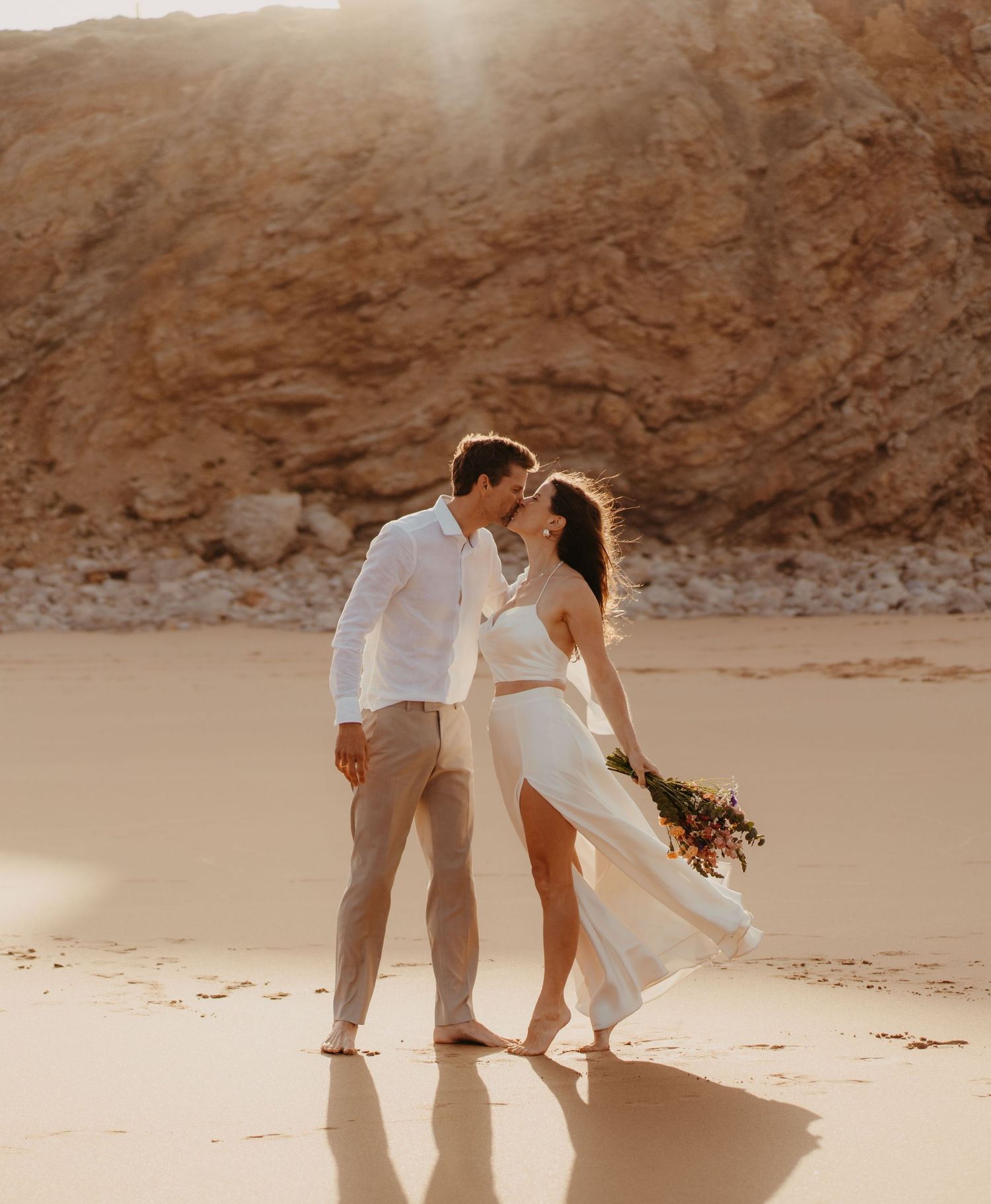 Bride and groom kissing barefoot on golden sandy beach with dramatic cliffs behind.