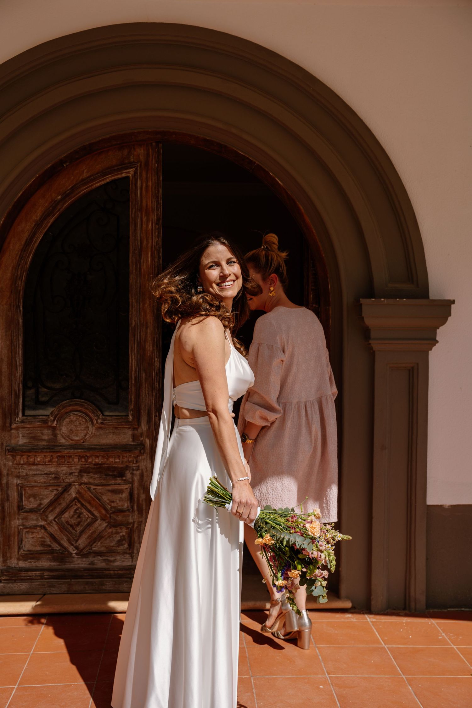 Smiling bride in white dress holding bouquet at ornate wooden church doorway.