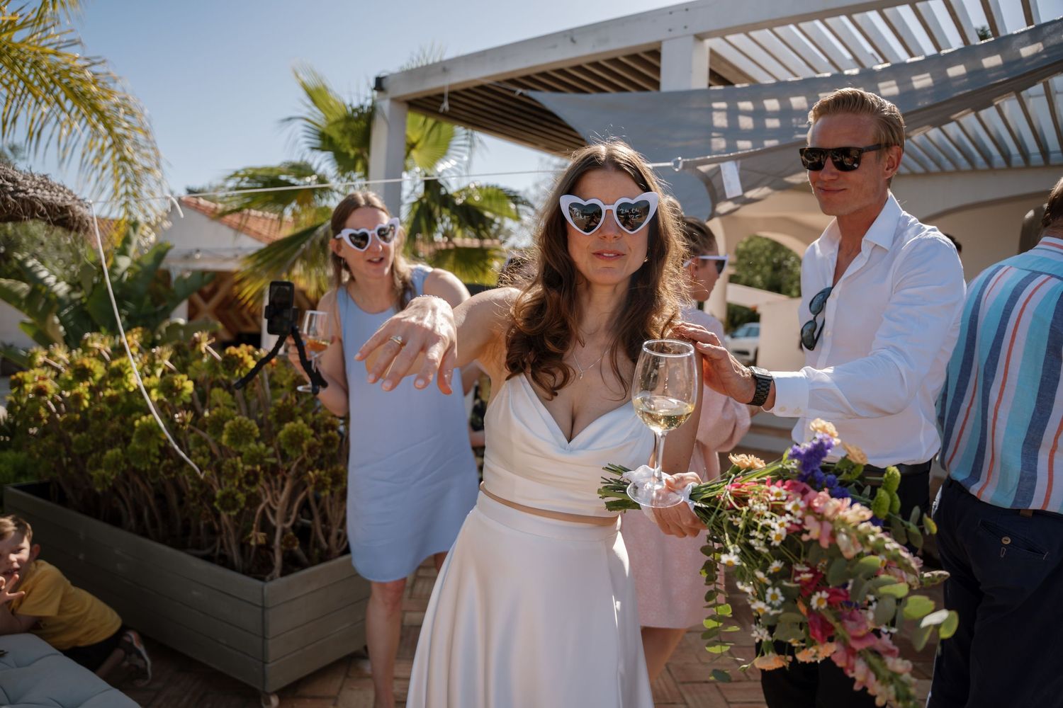 Bride in white dress with heart sunglasses holding champagne glass at outdoor wedding celebration.