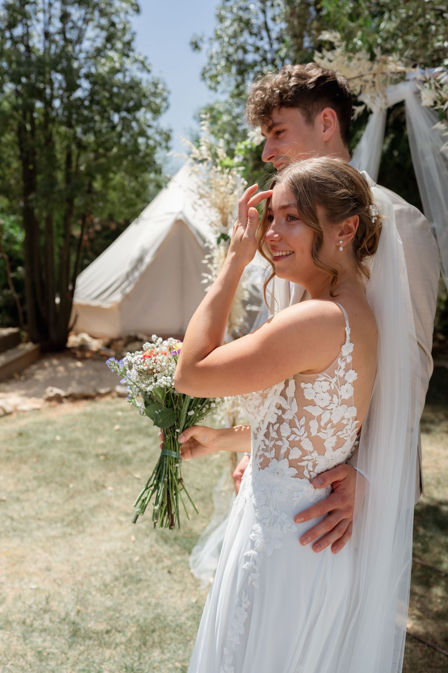 Bride in lace wedding dress holding wildflower bouquet embraced by groom outdoors near glamping tent.