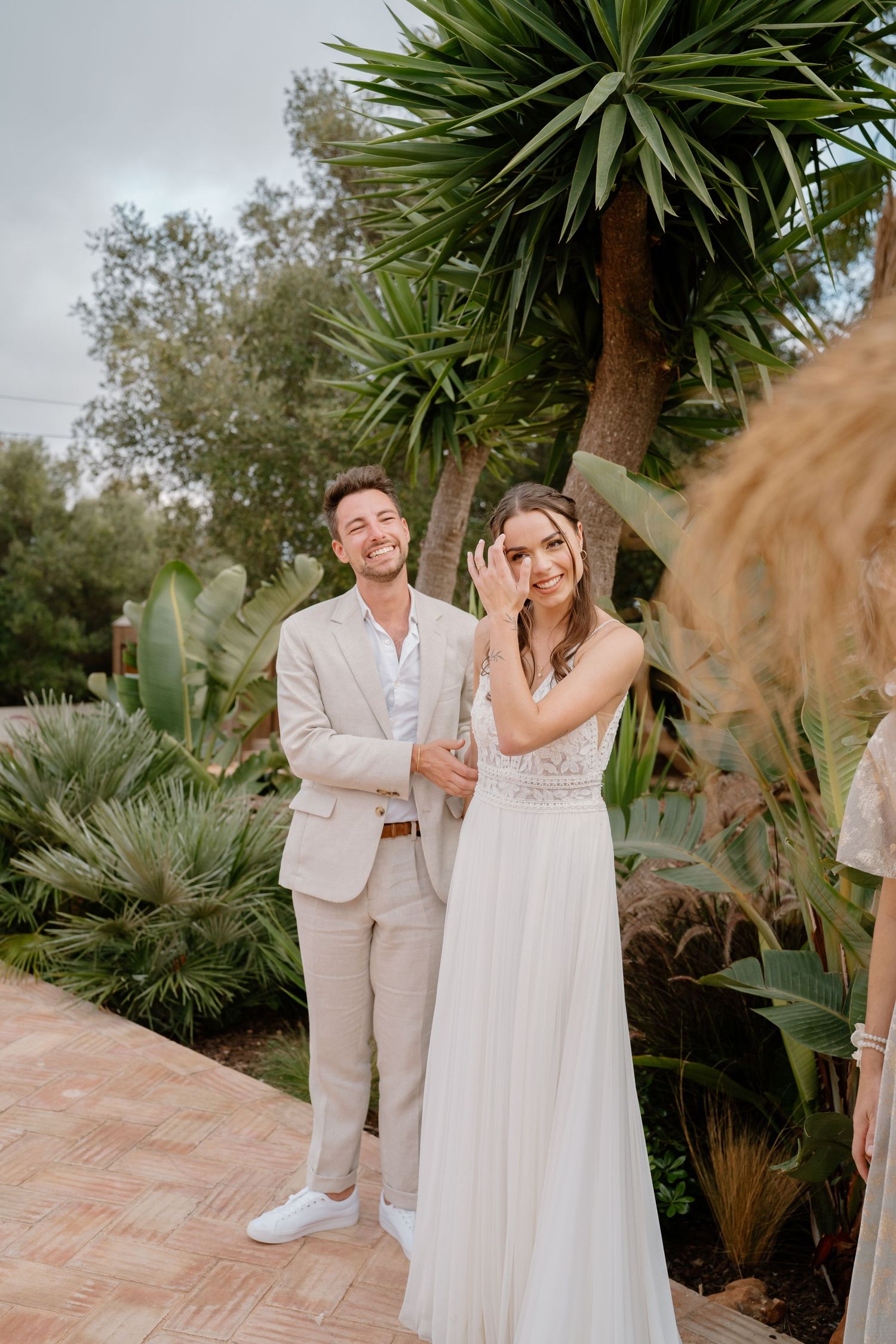 Smiling bride and groom in beige suit pose together outdoors surrounded by tropical plants at a garden wedding.