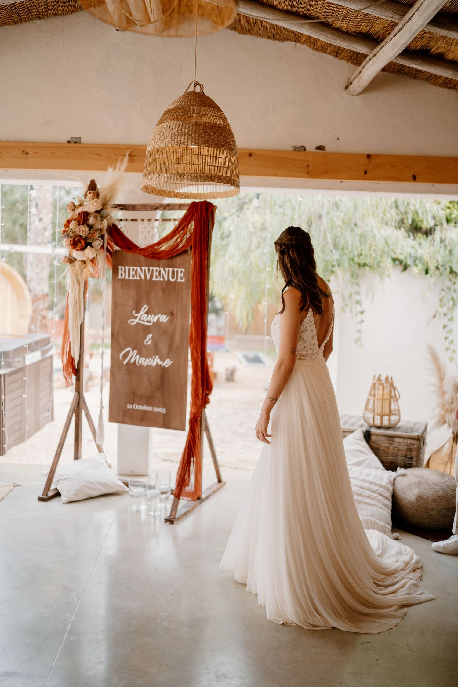 Bride in white gown stands before a rustic welcome sign reading Bienvenue at a boho wedding venue.