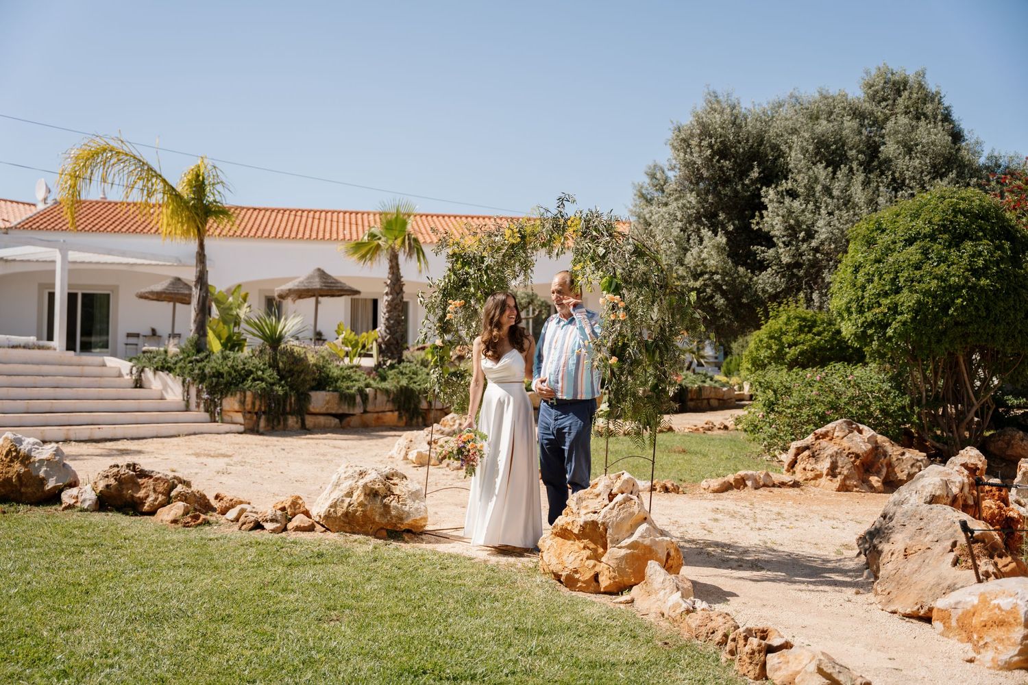 Bride and groom stroll through a sunny garden with rocky landscape beside a white Mediterranean villa.