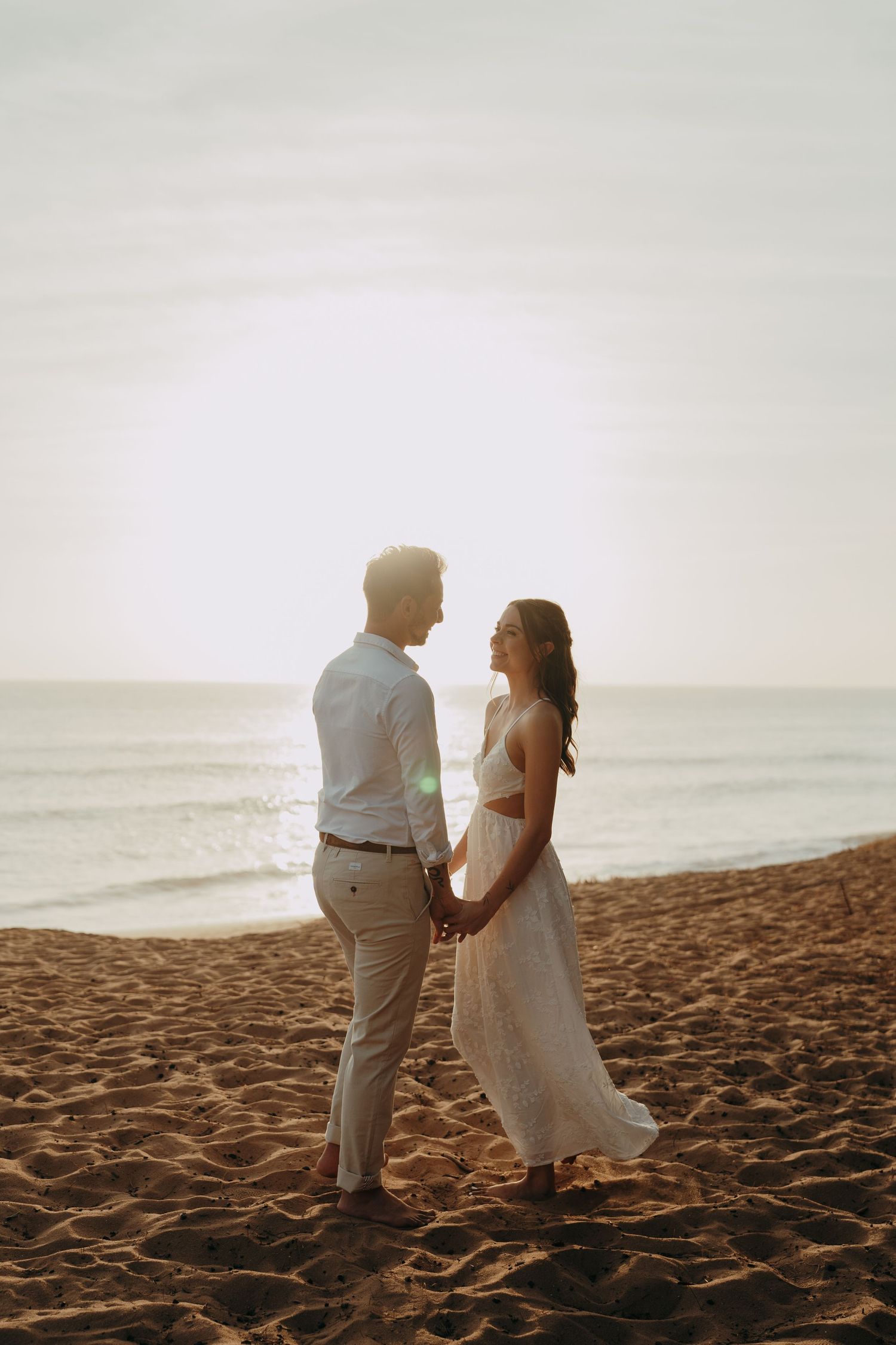 Couple holding hands on a sandy beach at golden sunset, woman in white dress, man in light blue shirt, ocean backdrop.