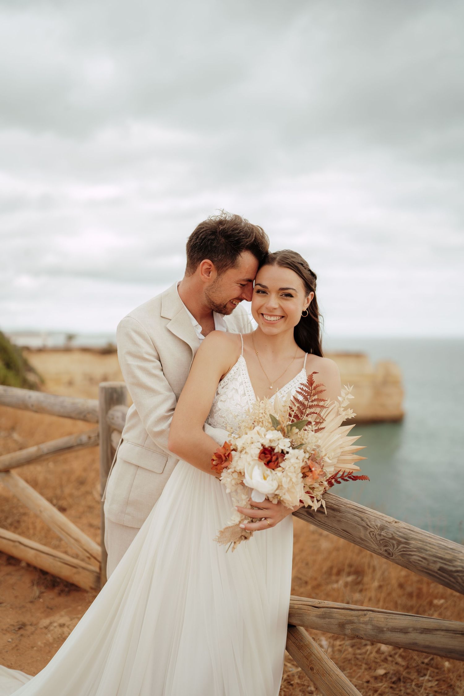 Bride and groom embracing on coastal cliff, bride holding boho bouquet of dried flowers, ocean backdrop.