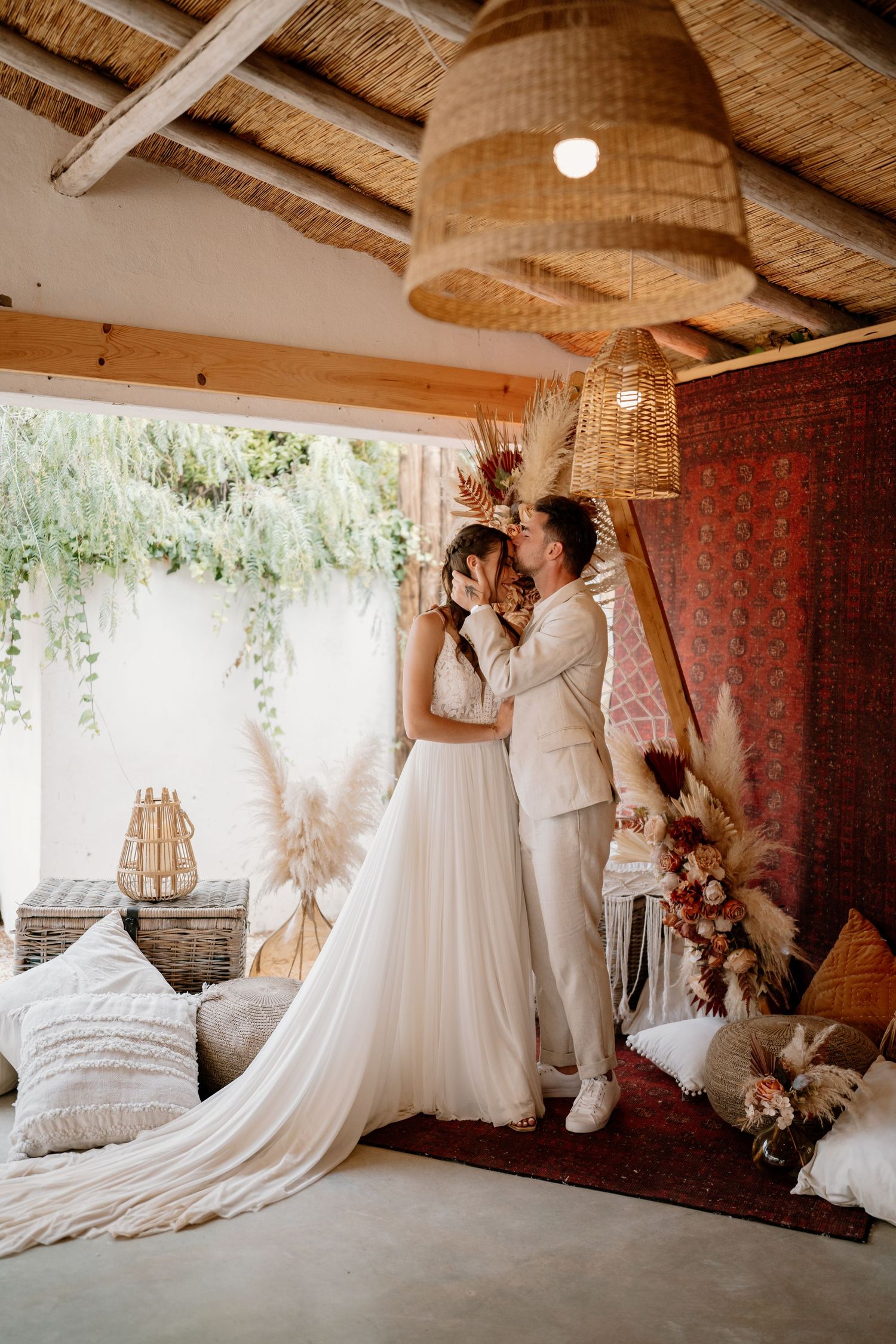 Bride and groom sharing a romantic kiss in a boho-styled wedding venue with pampas grass and rattan decor.