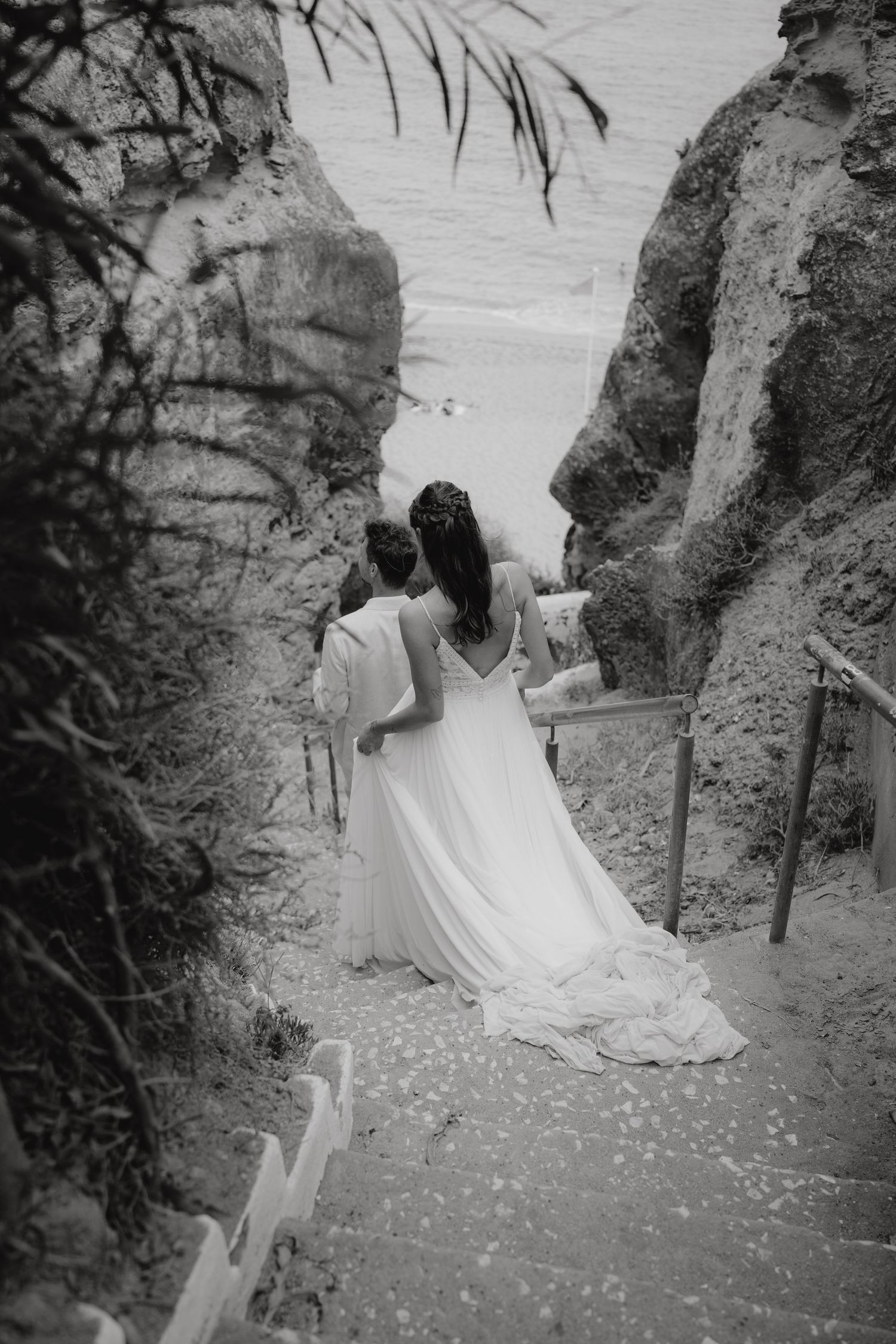 Black and white photo of two brides in white gowns walking down rocky coastal stairs toward the ocean.