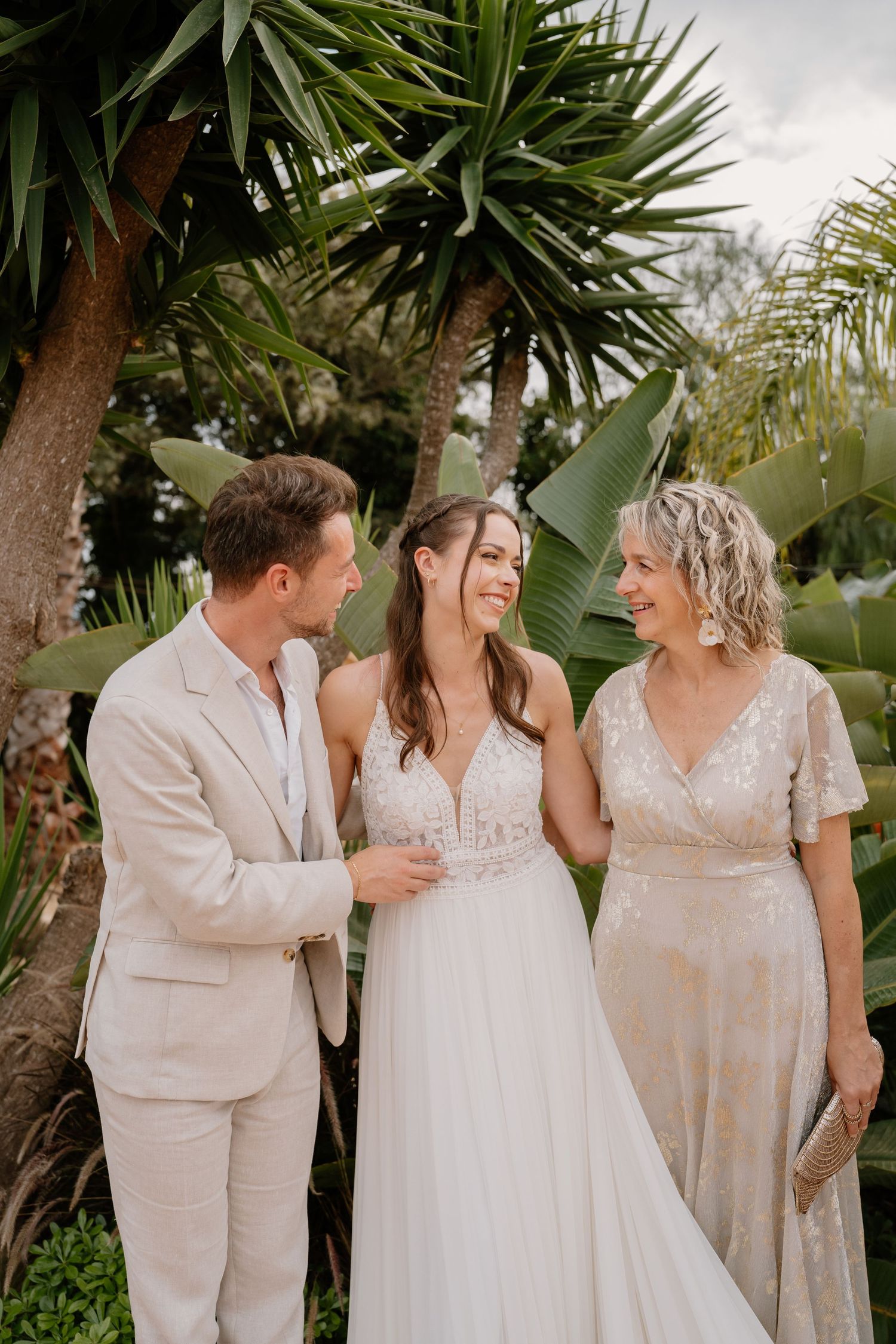 Bride in white gown smiles with groom in beige suit and guest in sequin dress amid tropical palms.
