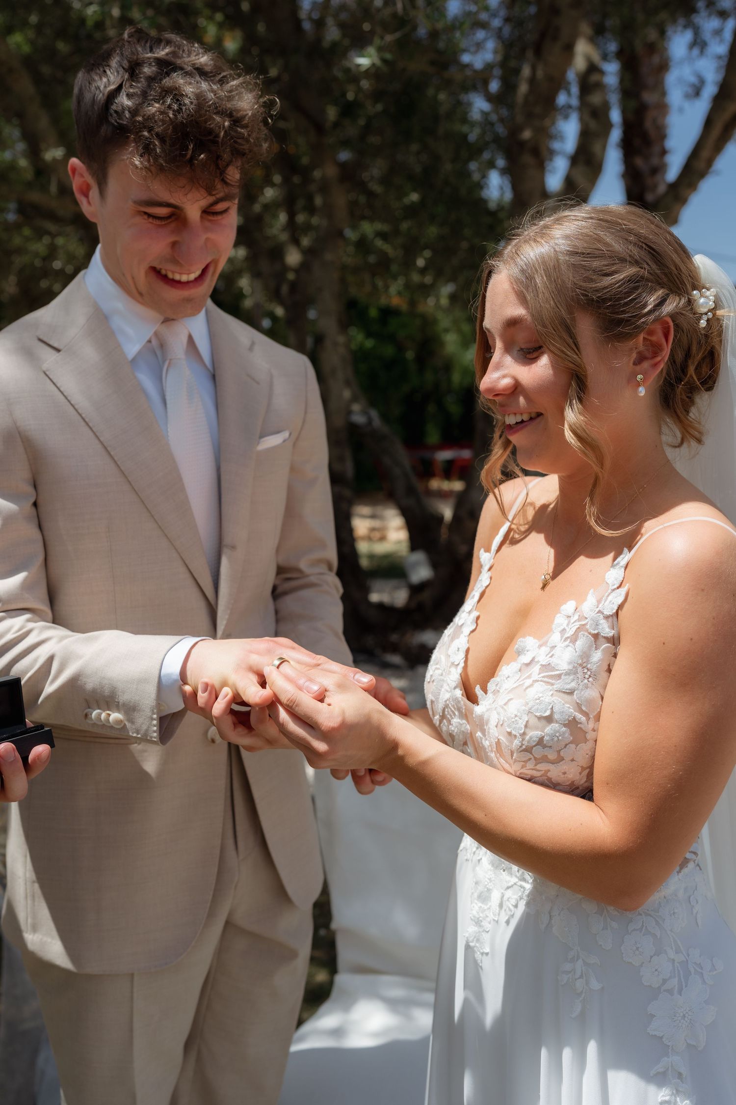 Groom in beige suit placing wedding ring on bride's finger during outdoor ceremony.