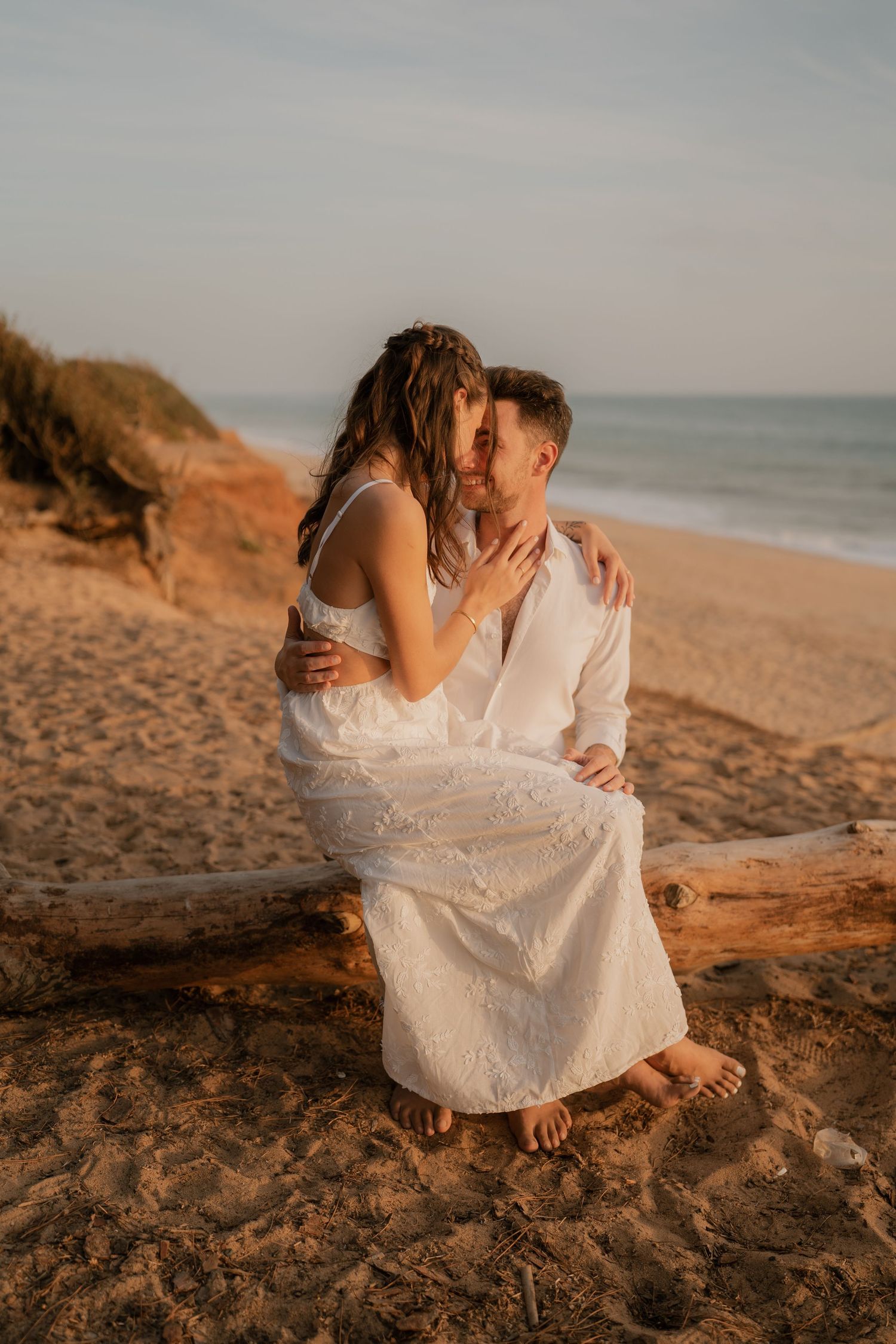 Couple in white embracing on driftwood at golden hour beach, sandy dunes and ocean in background.