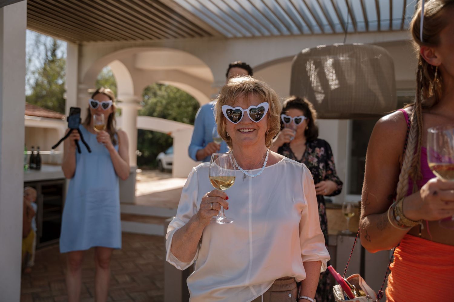 Woman in white blouse smiling and holding champagne glass at sunny outdoor wedding reception.