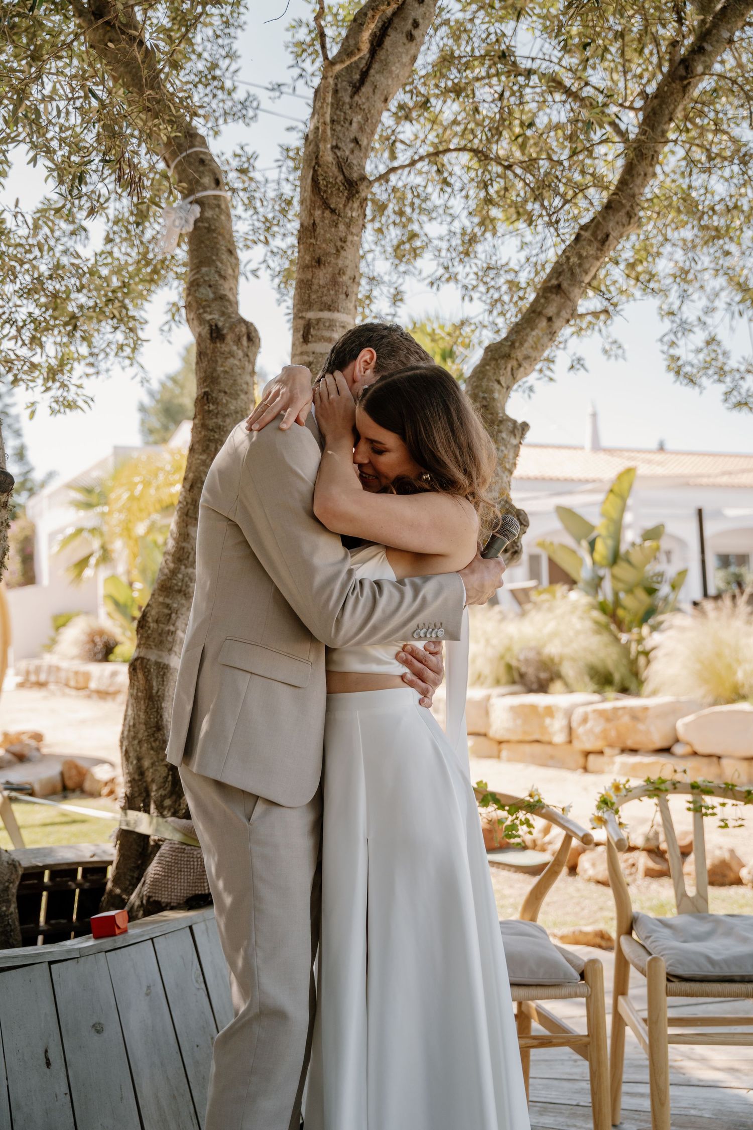 Newlyweds share an emotional hug beneath olive trees at a sunlit outdoor wedding ceremony.