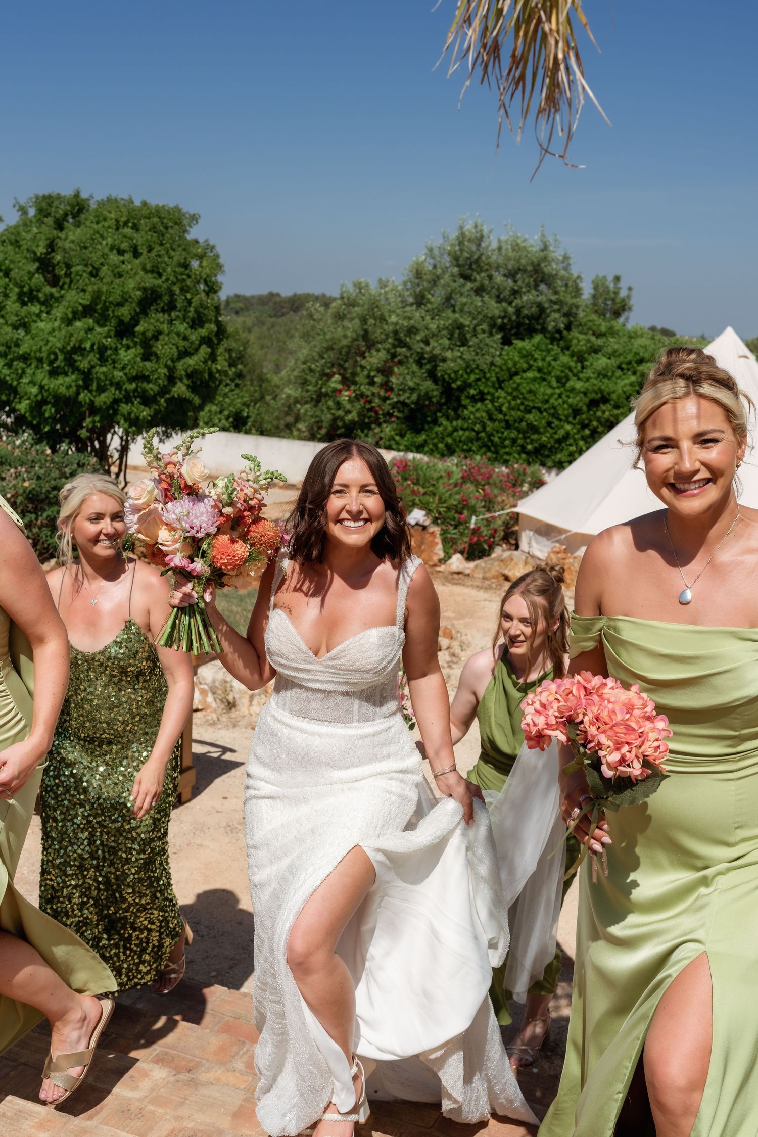 Bride in white gown walks with smiling bridesmaids in green dresses holding colorful bouquets outdoors on a sunny day.