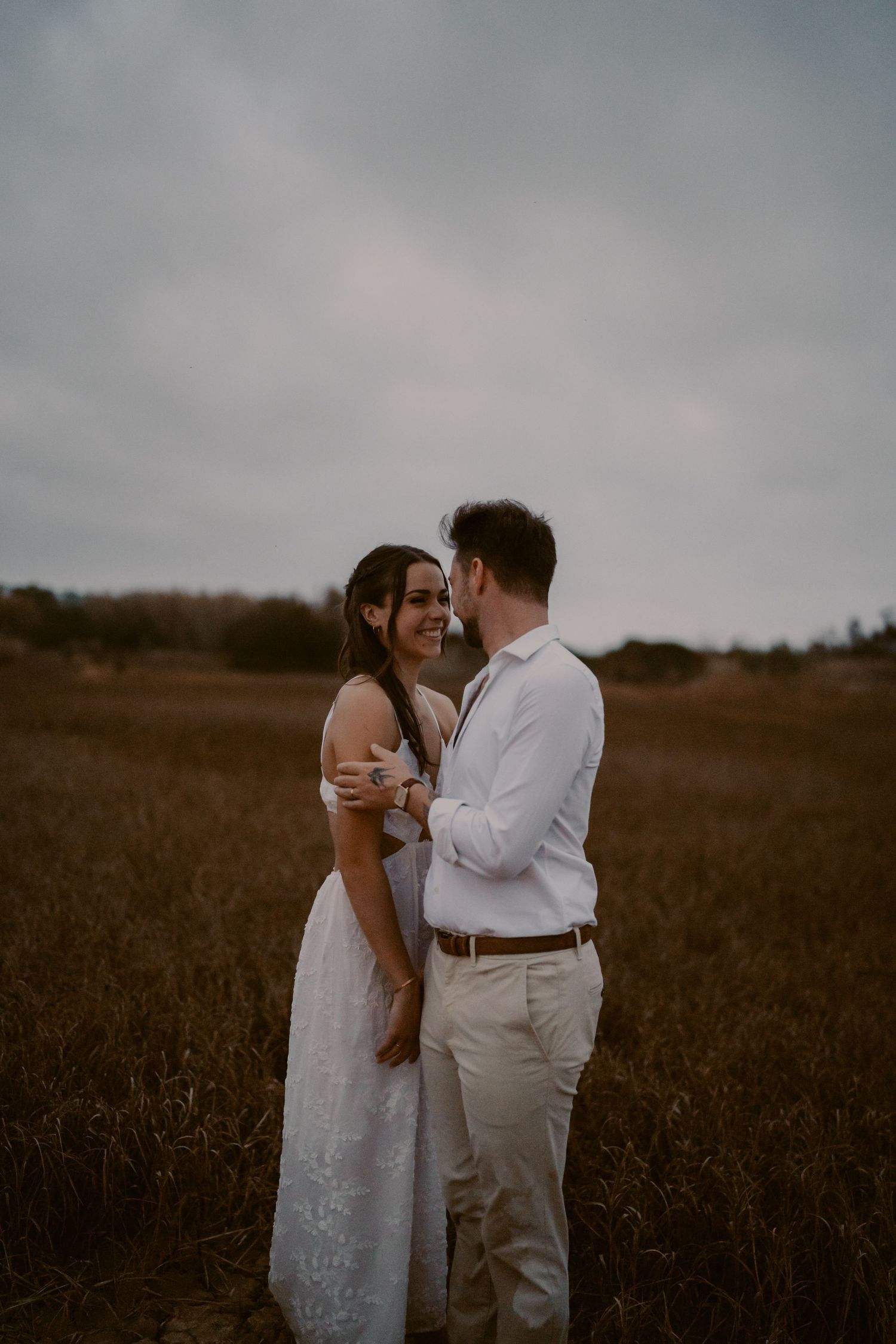 Couple embracing in a golden field, woman in white dress smiling at man in white shirt, moody sky backdrop.