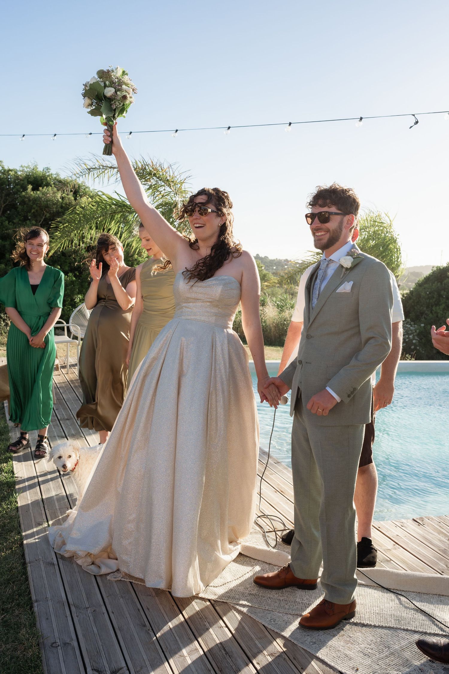 Joyful bride raises bouquet while holding hands with groom in gray suit beside outdoor pool at sunny wedding ceremony.