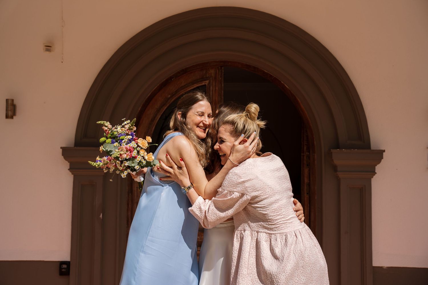 Two women sharing a joyful embrace outside an arched church entrance with bouquet.