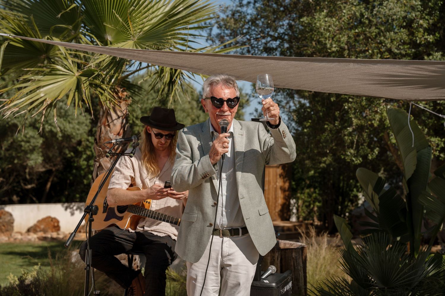 Man in grey suit giving speech with microphone while guitarist performs at outdoor wedding ceremony.
