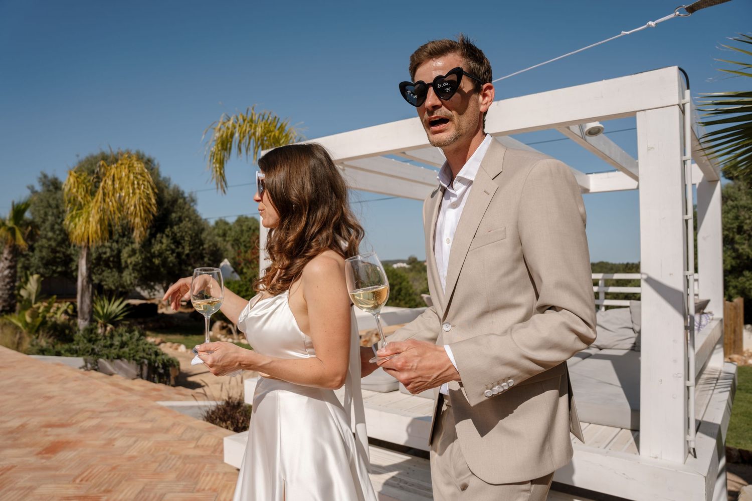 Couple in wedding attire holding champagne glasses standing at sunny outdoor wedding reception.