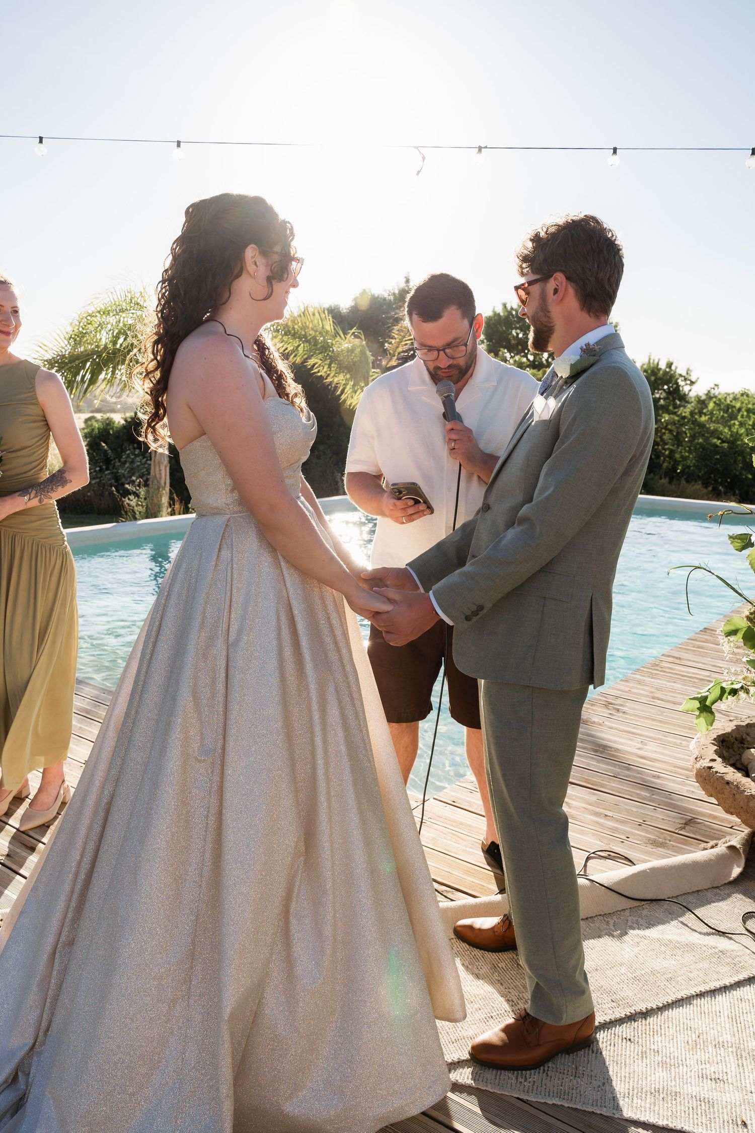 Bride and groom exchange vows poolside at outdoor wedding ceremony, officiant reads from notes in sunny backyard setting.