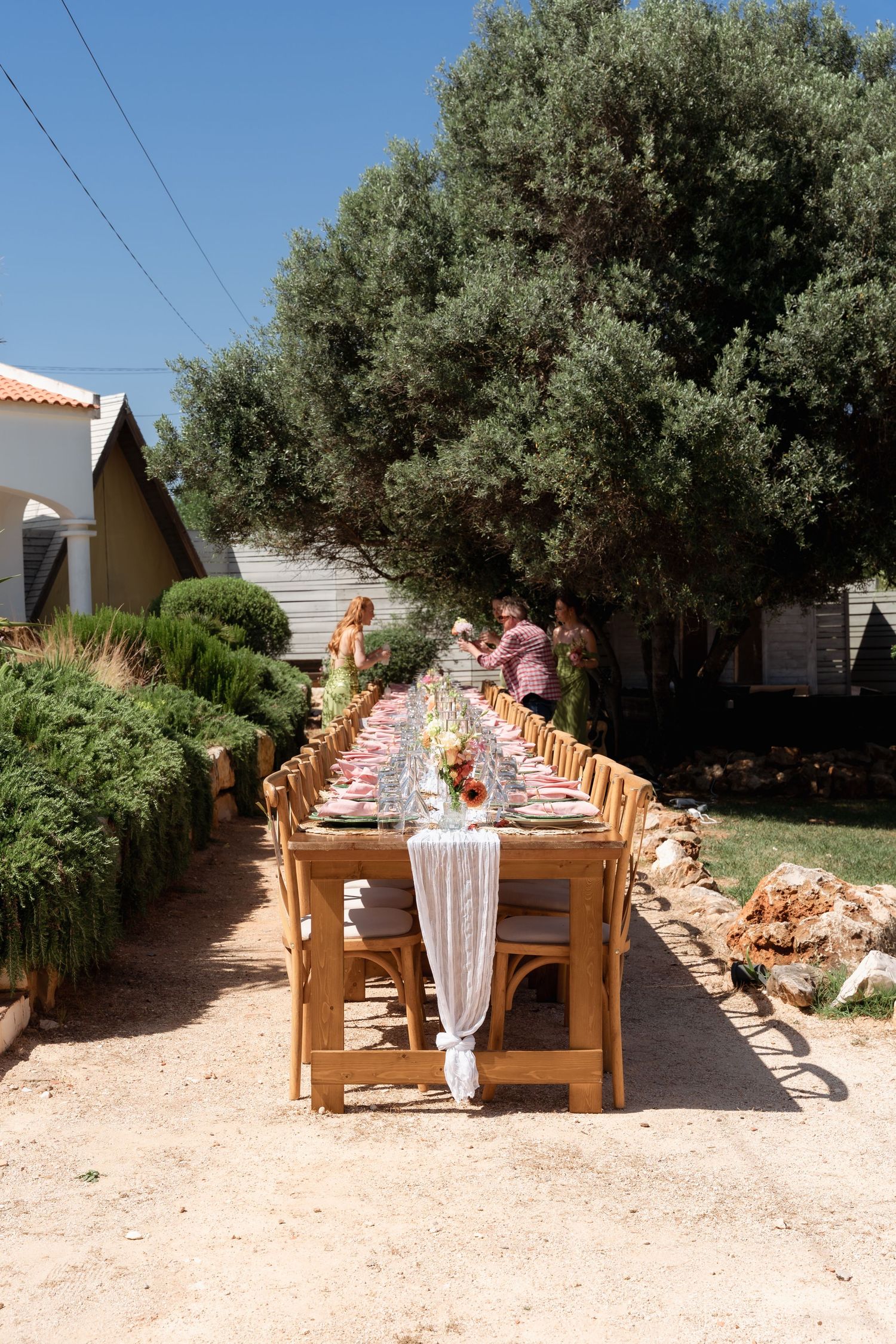 Long outdoor dining table set for a garden party with wooden chairs, floral centerpieces, and lush greenery under a clear blue sky.