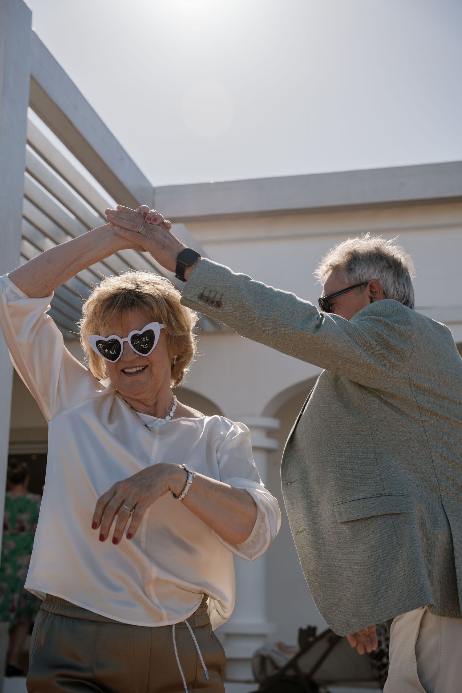 Senior couple dancing joyfully outdoors at a wedding, woman wearing heart-shaped sunglasses.