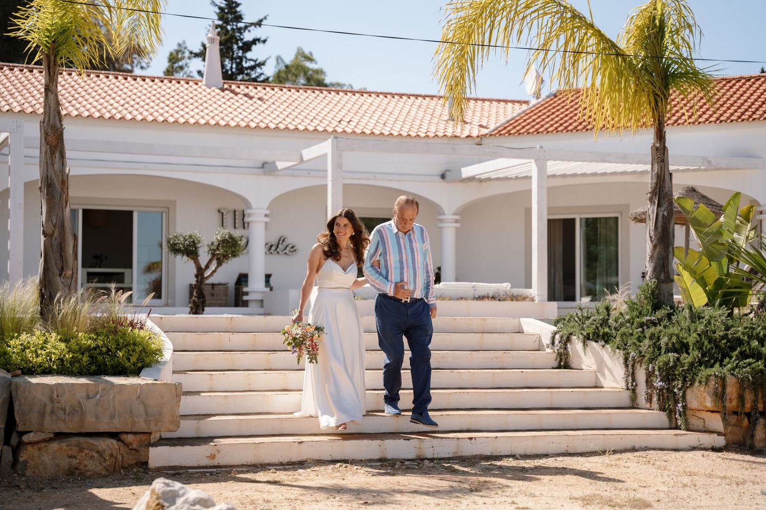 Bride and groom walk down stone steps at a white Mediterranean villa wedding venue on a sunny day.