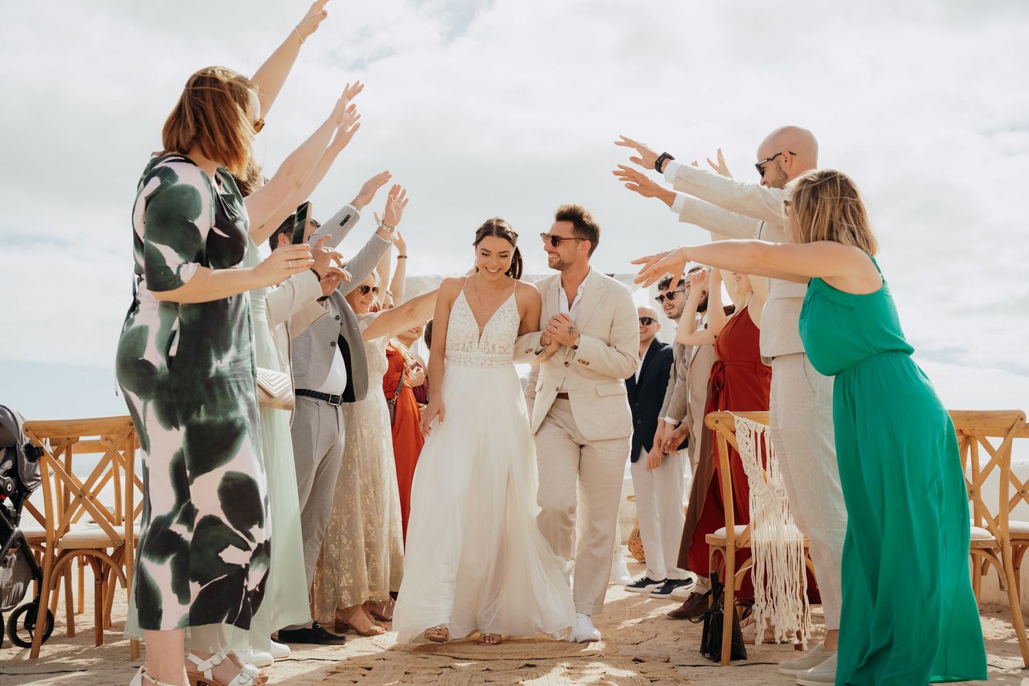 Bride and groom walk through cheering guests at outdoor beach wedding ceremony under blue sky.