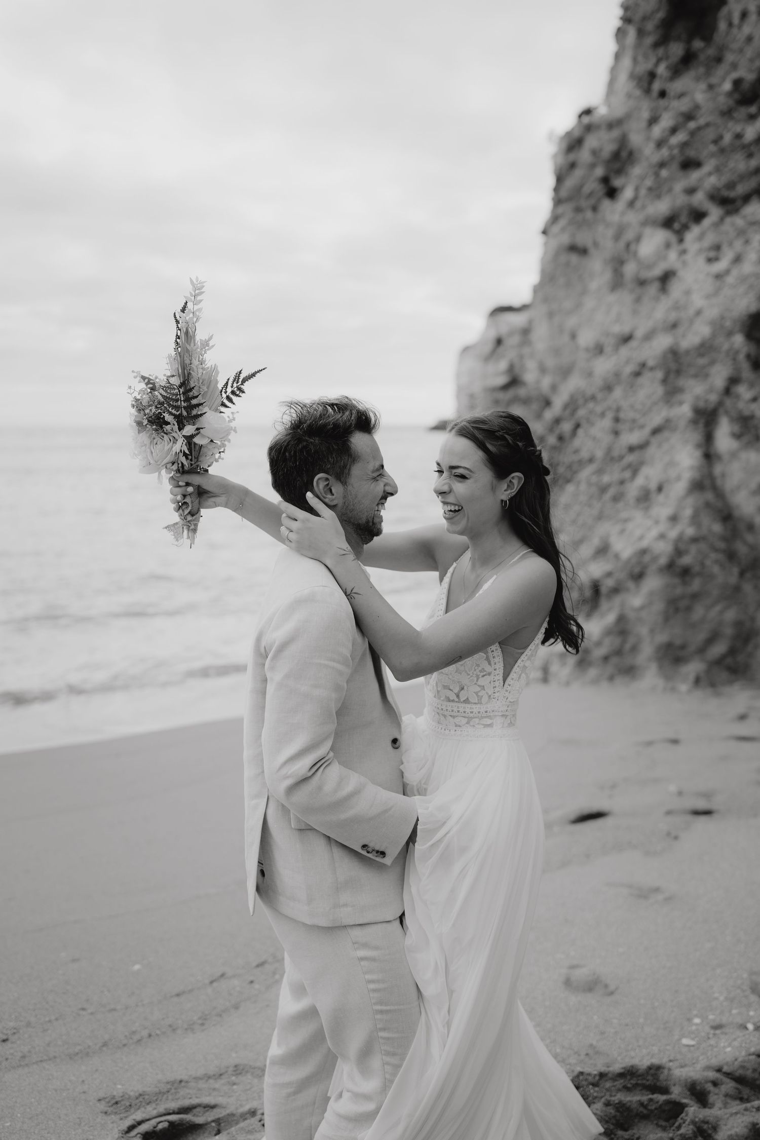 Black and white photo of a couple in wedding attire embracing on a beach, bride holding a bouquet near rocky cliffs.