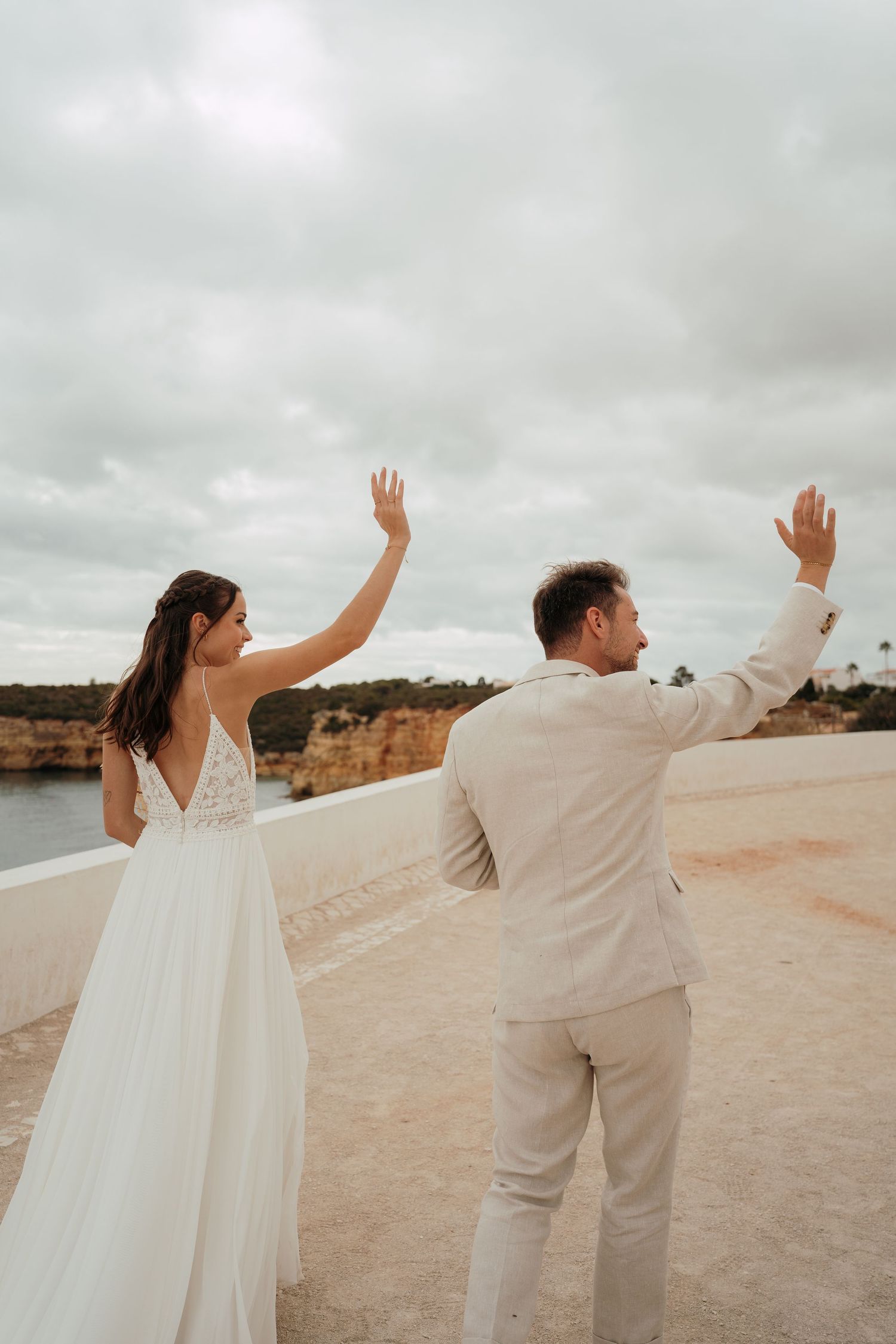 Bride and groom waving hands together on a coastal cliff during their beach wedding in the Algarve, Portugal.