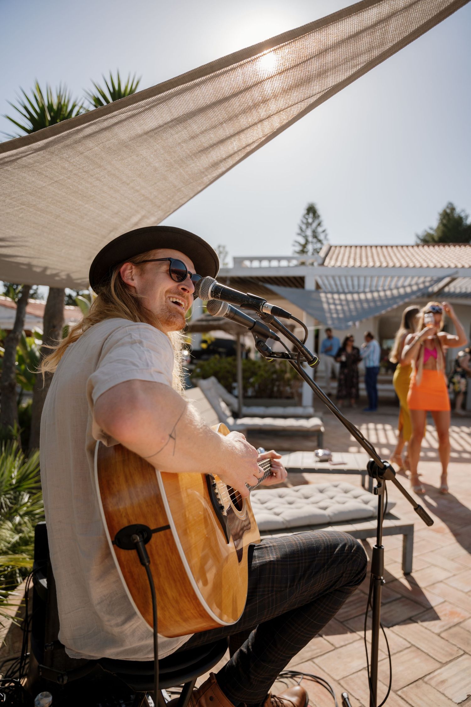 Long-haired guitarist in a hat performs outdoors at a sunny poolside wedding reception.