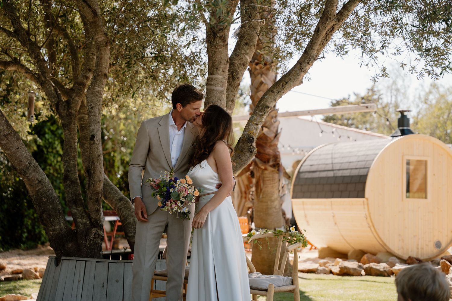 Bride and groom share a kiss holding a colorful bouquet under olive trees at outdoor wedding.